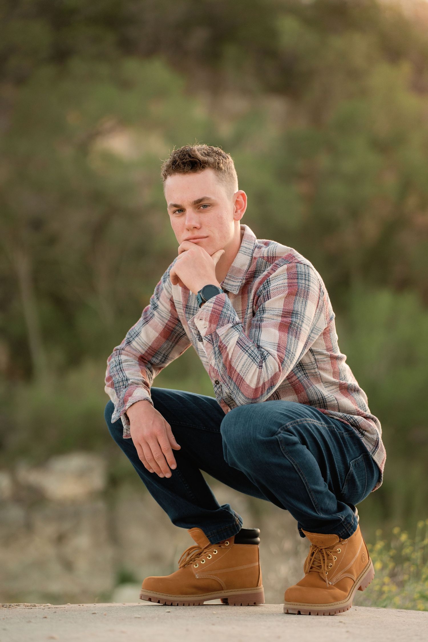 Young man in plaid shirt and tan boots crouching outdoors with hand on chin in a thoughtful pose.