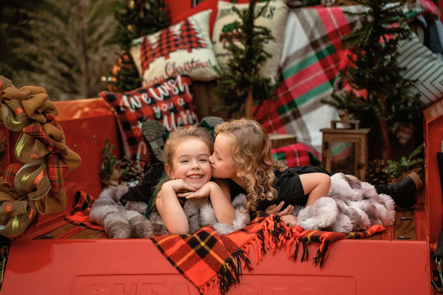 Two young girls share a sweet kiss in a festive red truck surrounded by Christmas decorations and plaid holiday accents.