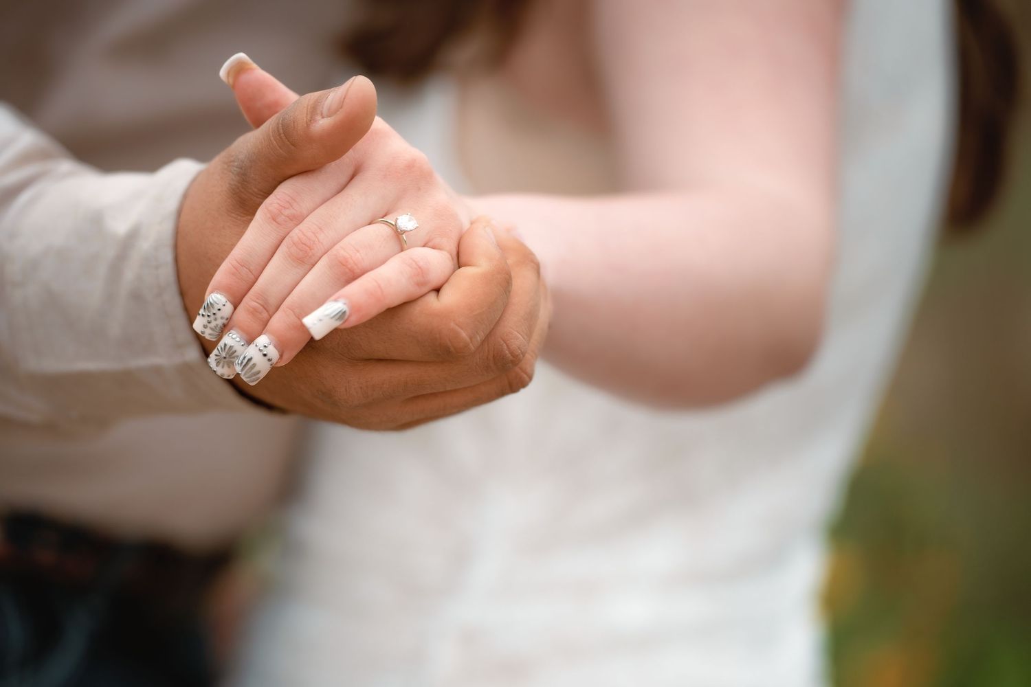 Close up of hands holding each other with an engagement ring in focus during a romantic moment outdoors.