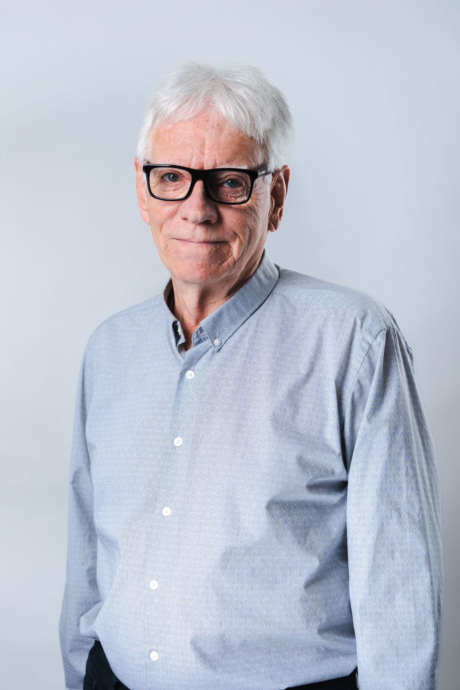 Professional studio portrait of a senior person wearing glasses and a light gray button-down shirt against a plain background.