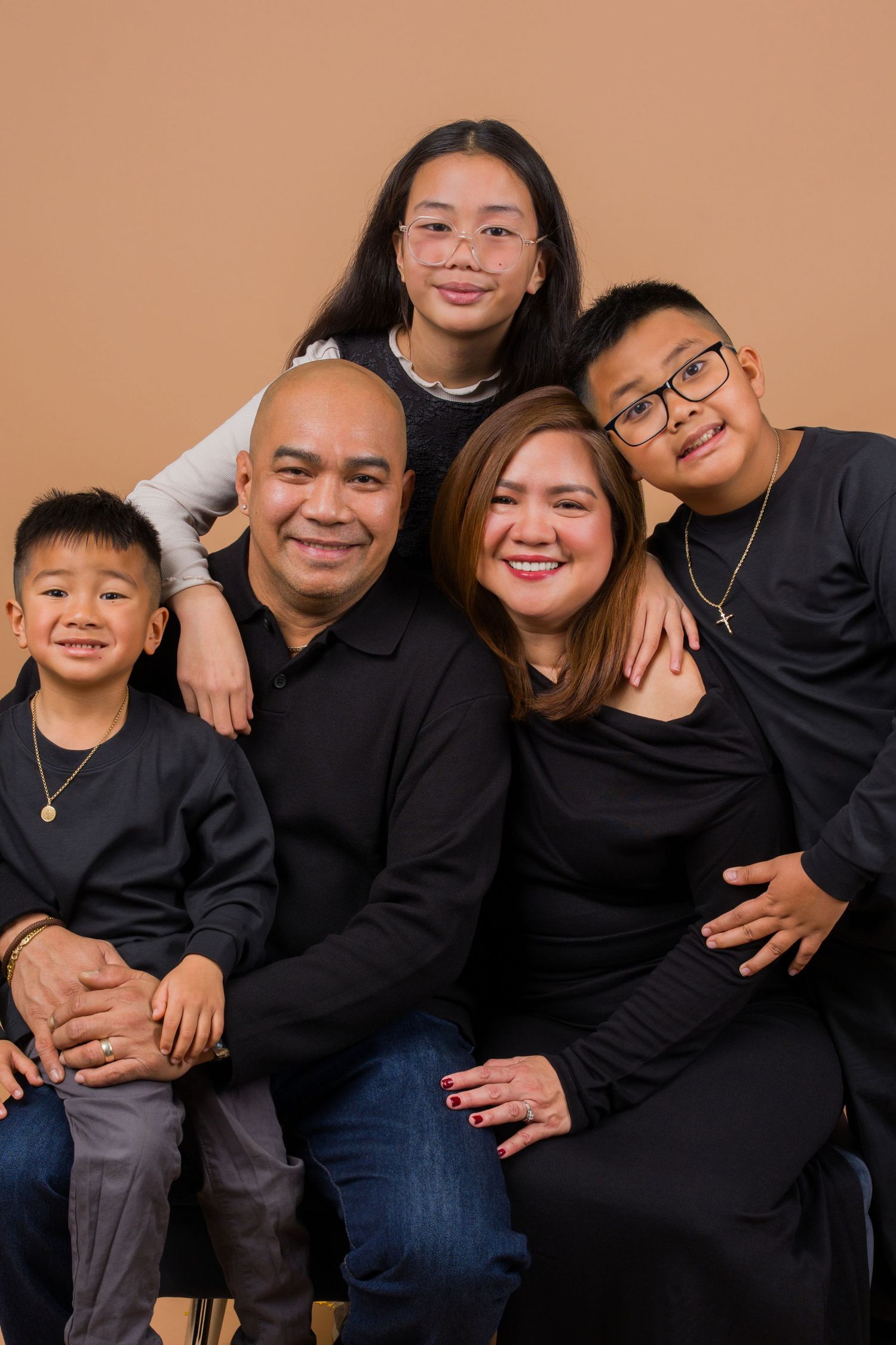 Family wearing matching black outfits poses together for casual portrait session on brown backdrop.