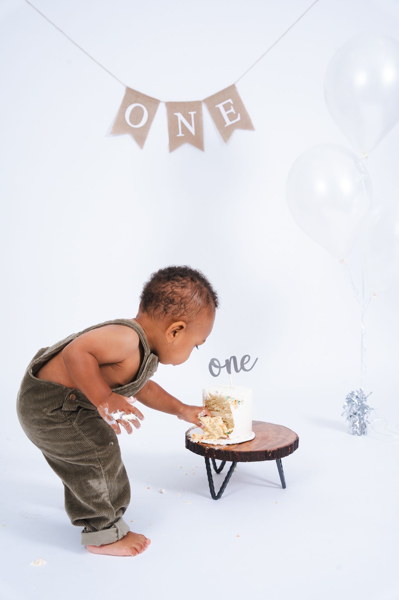 Baby in brown overalls eagerly dives into a smash cake during first birthday photoshoot with white backdrop and banner.