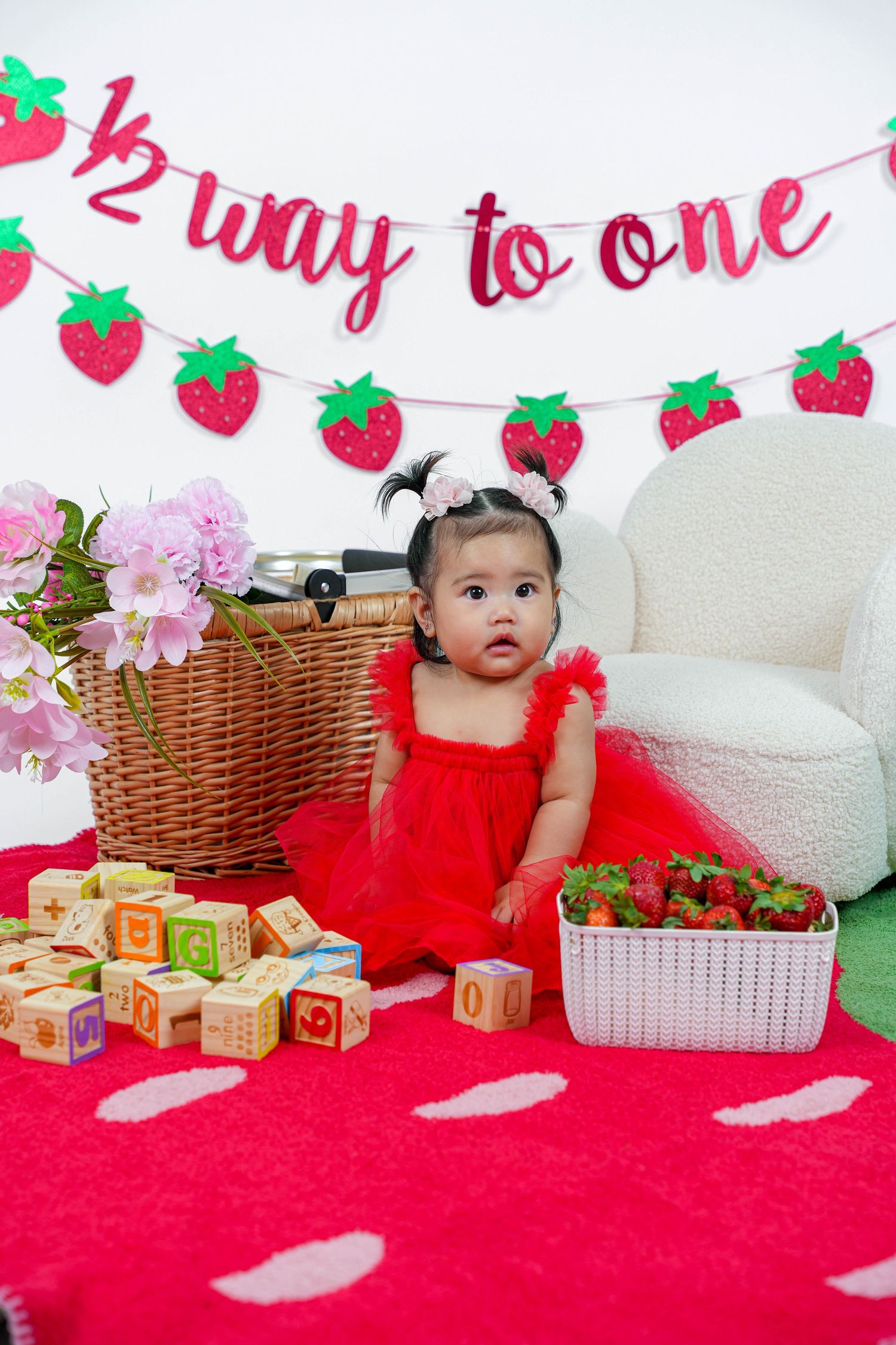 Baby in red outfit lies on pink and white polka dot backdrop with wooden blocks and strawberry themed party decorations.