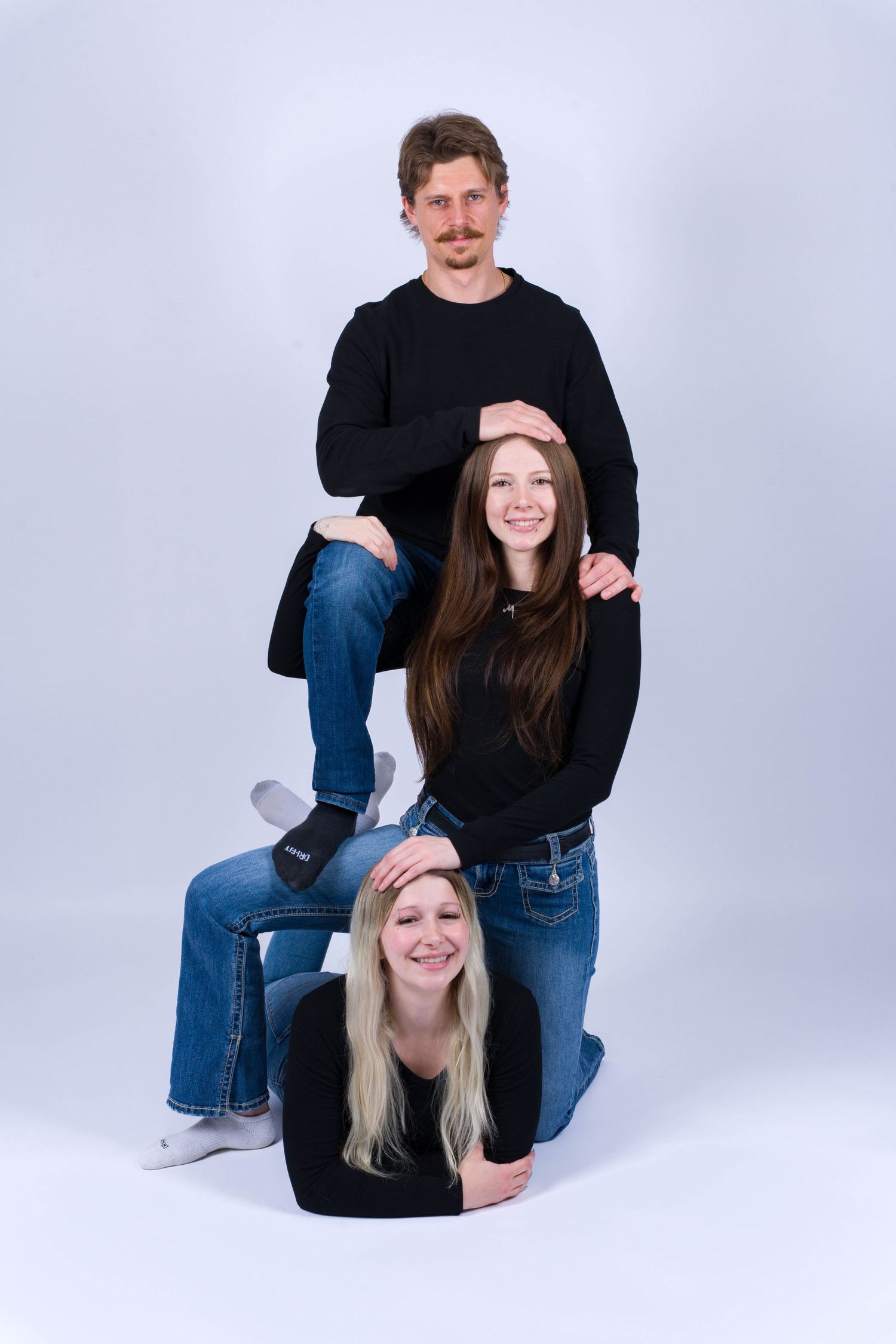 Three people in black sweaters pose creatively stacked on top of each other against a light gray studio background.