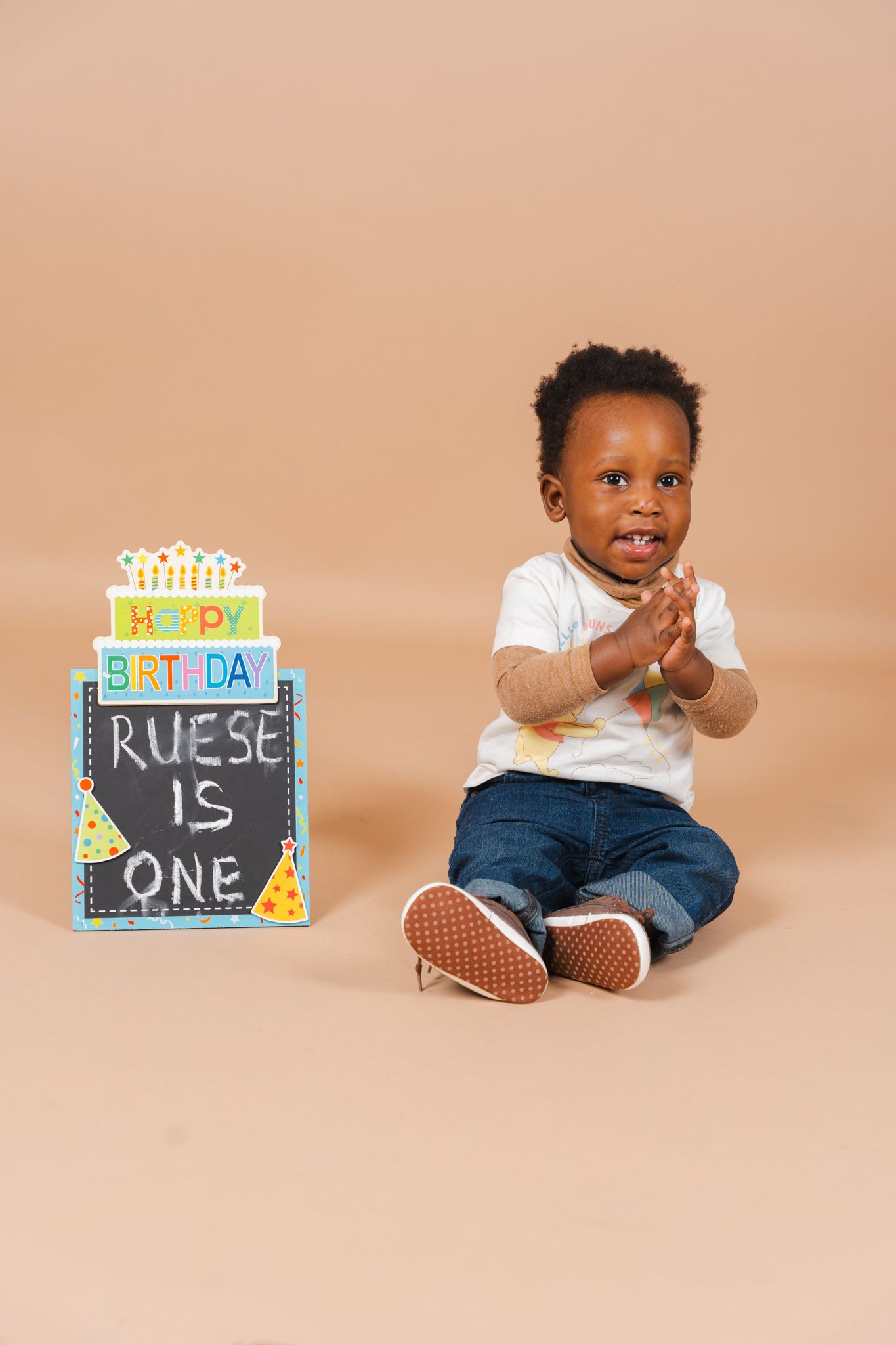 Baby wearing white shirt and jeans sits next to colorful first birthday sign on beige backdrop.