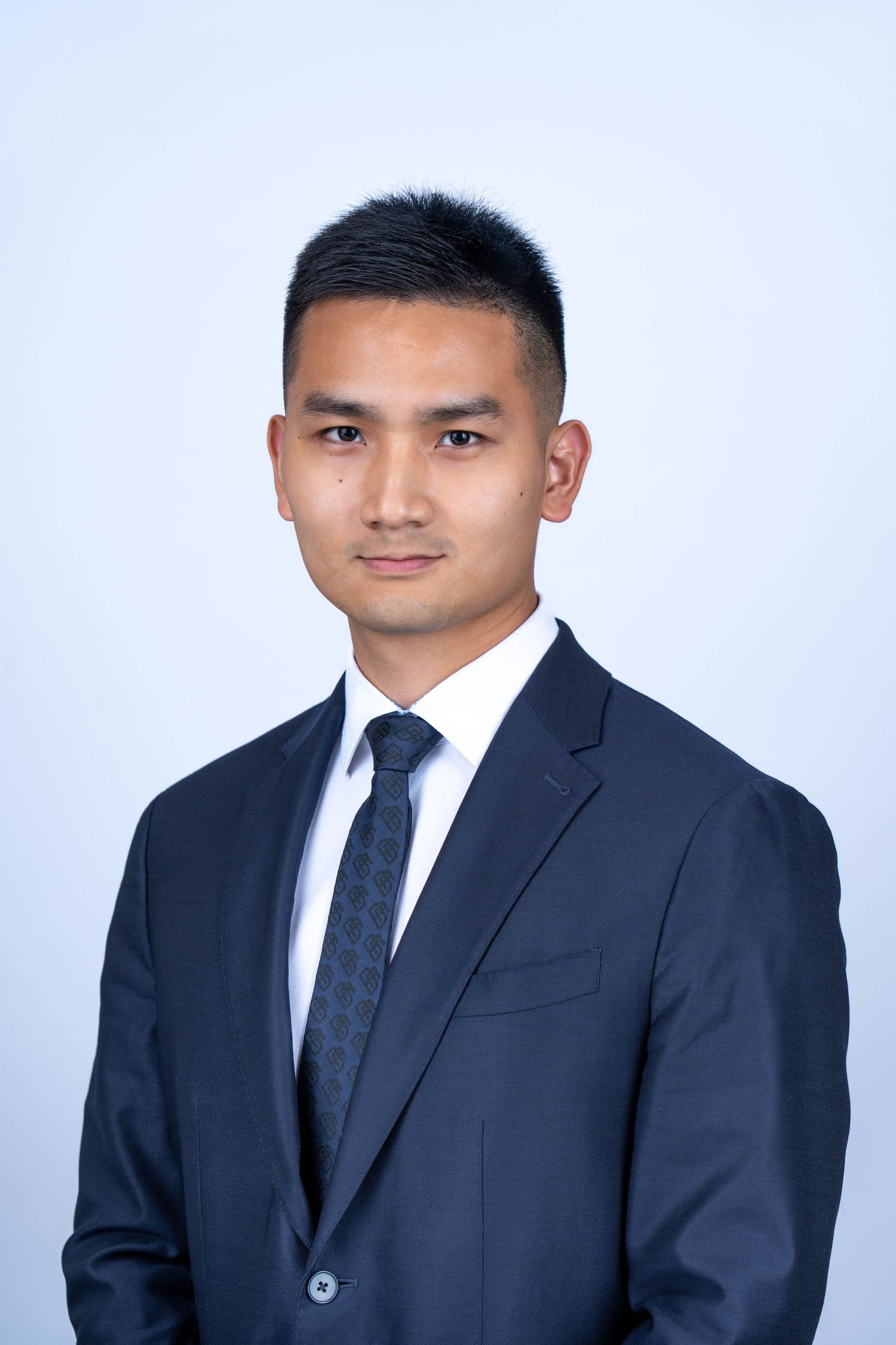 Professional headshot portrait of person in navy suit and tie against light blue background.