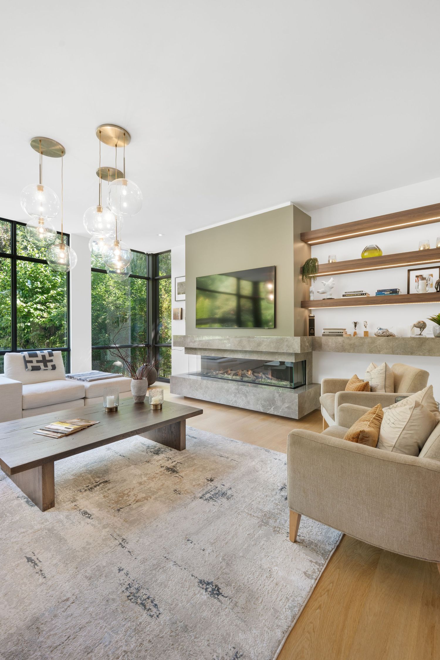 Modern living room with beige sofa, hardwood floors, and large windows overlooking green landscape.