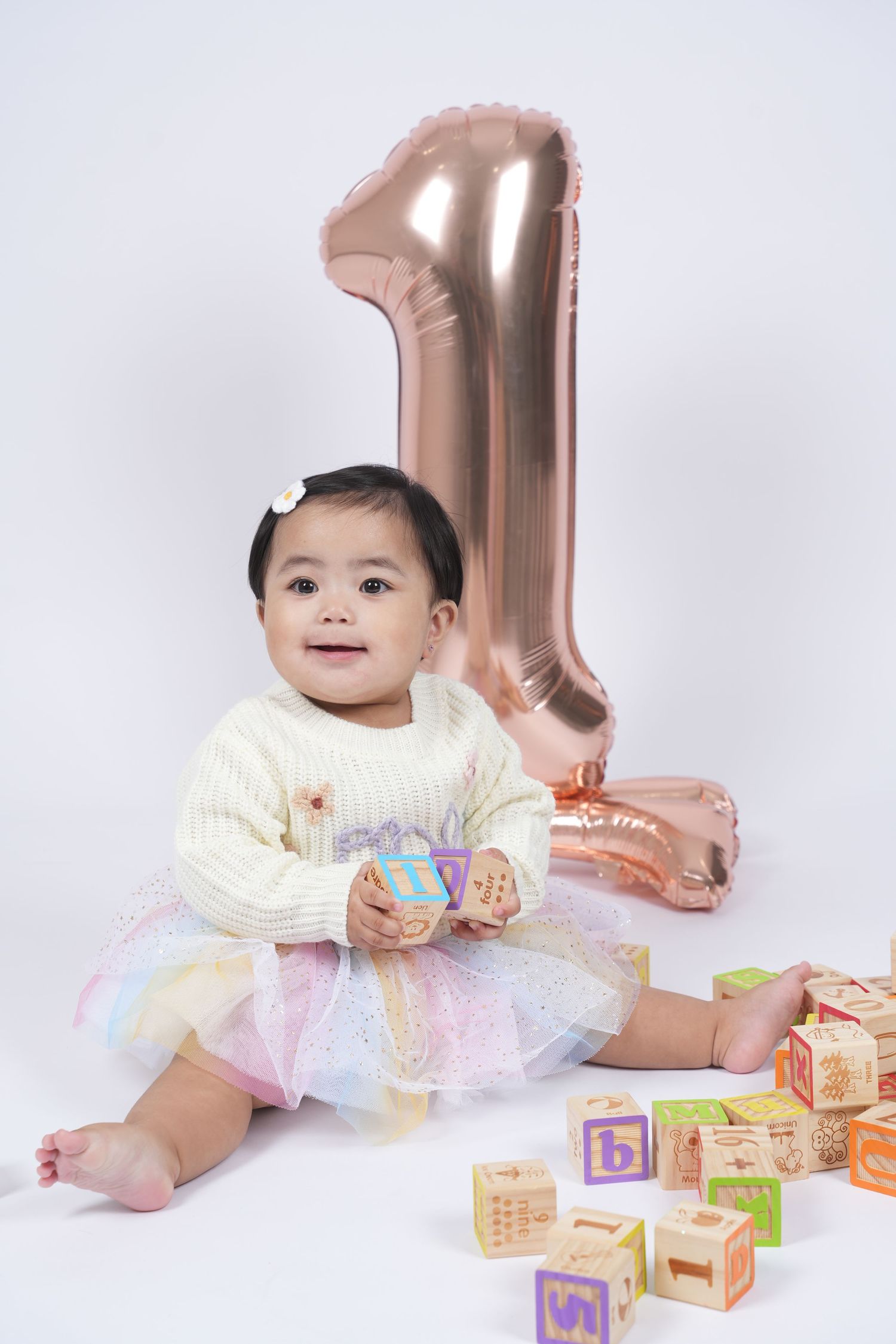 A baby in a white sweater and tulle skirt sits with alphabet blocks and a large rose gold number one balloon.
