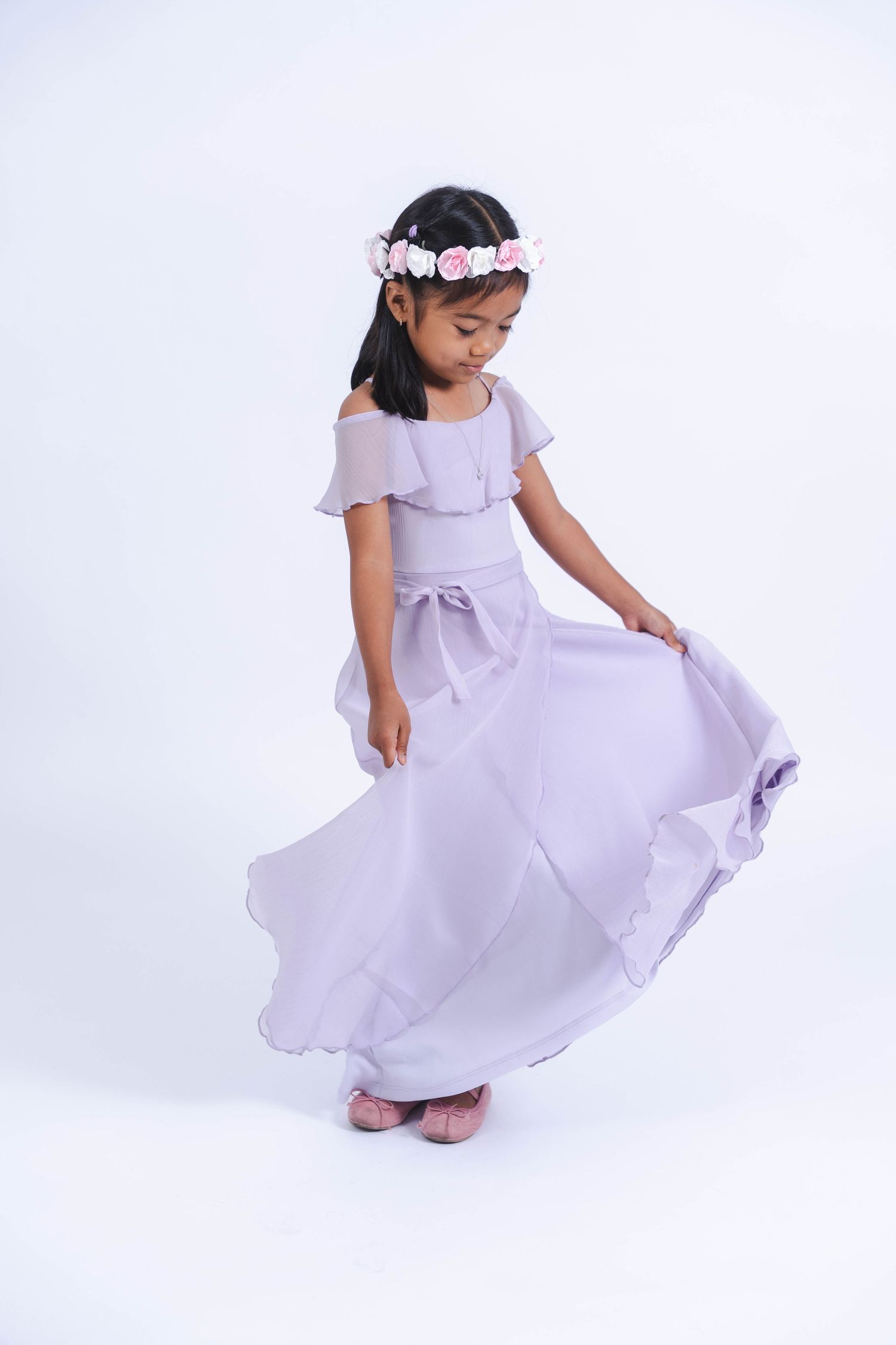 Young girl twirls in a flowing lavender dress with fluttering sleeves and bow detail against a white backdrop.