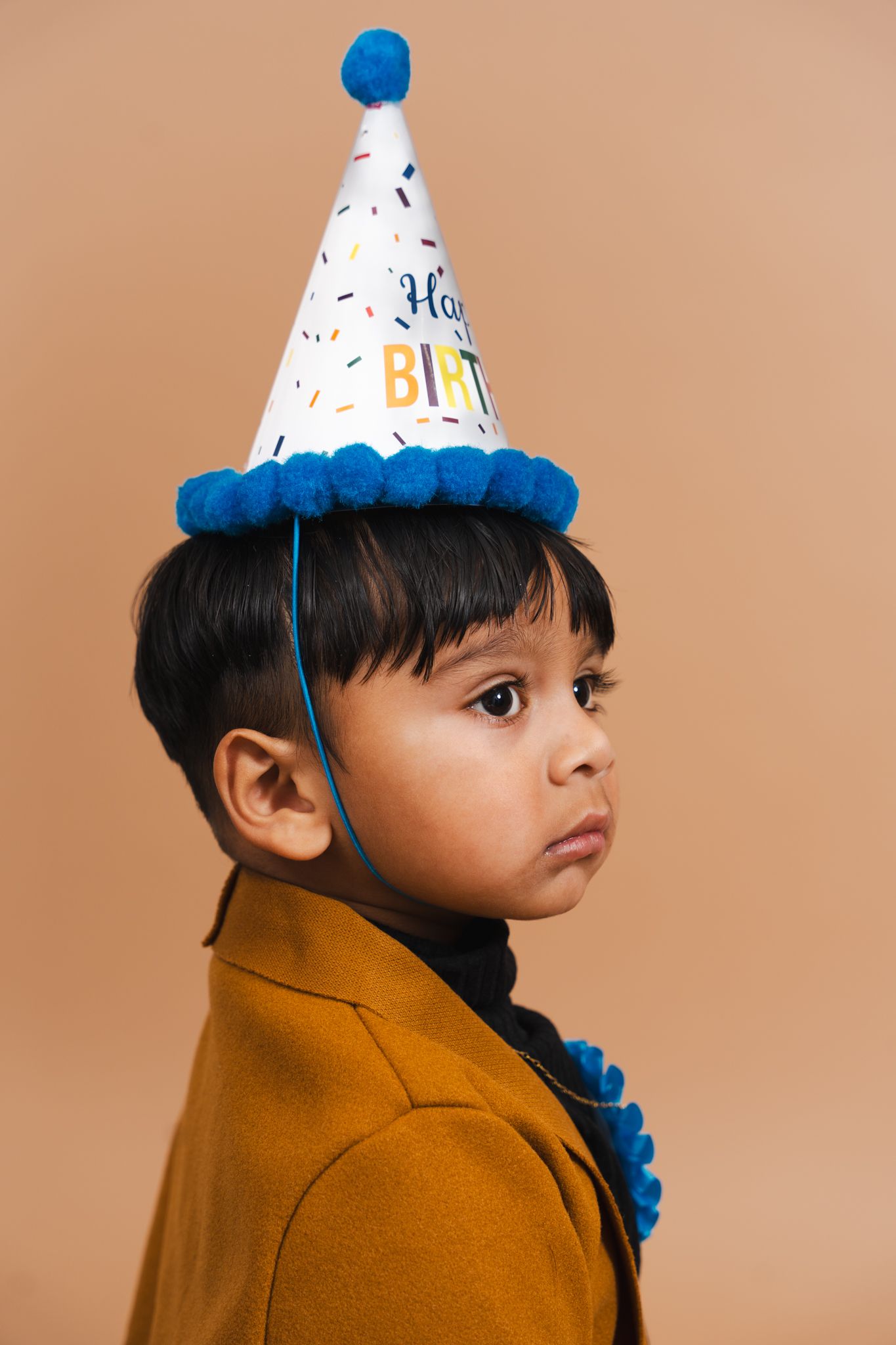 A side profile view of a child wearing a brown coat and birthday party hat against a neutral background.