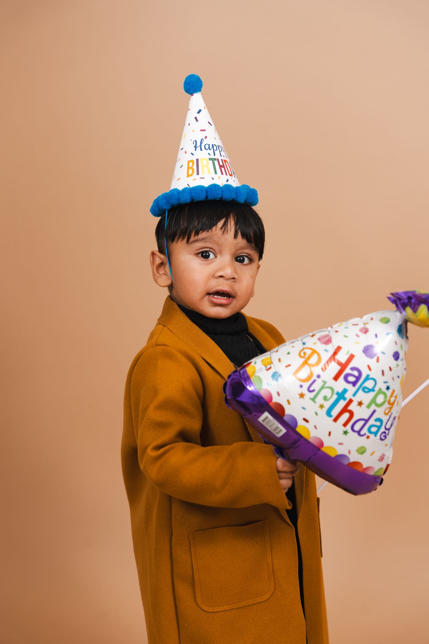A small child in a brown coat and party hat plays with a colorful birthday balloon against a beige backdrop.