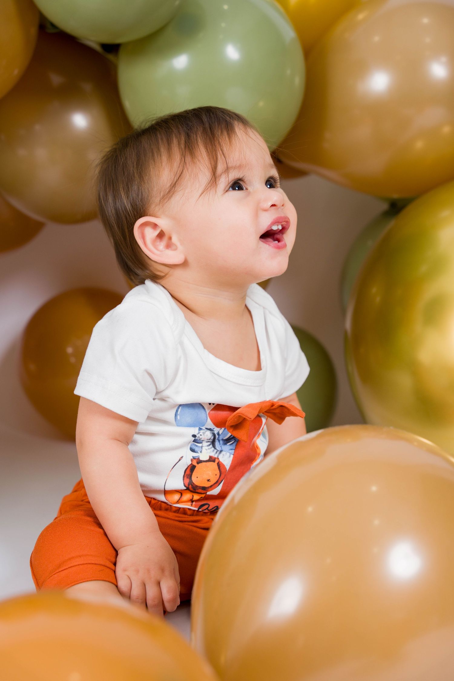 Baby in white t-shirt plays joyfully among gold and beige balloons at a festive celebration.