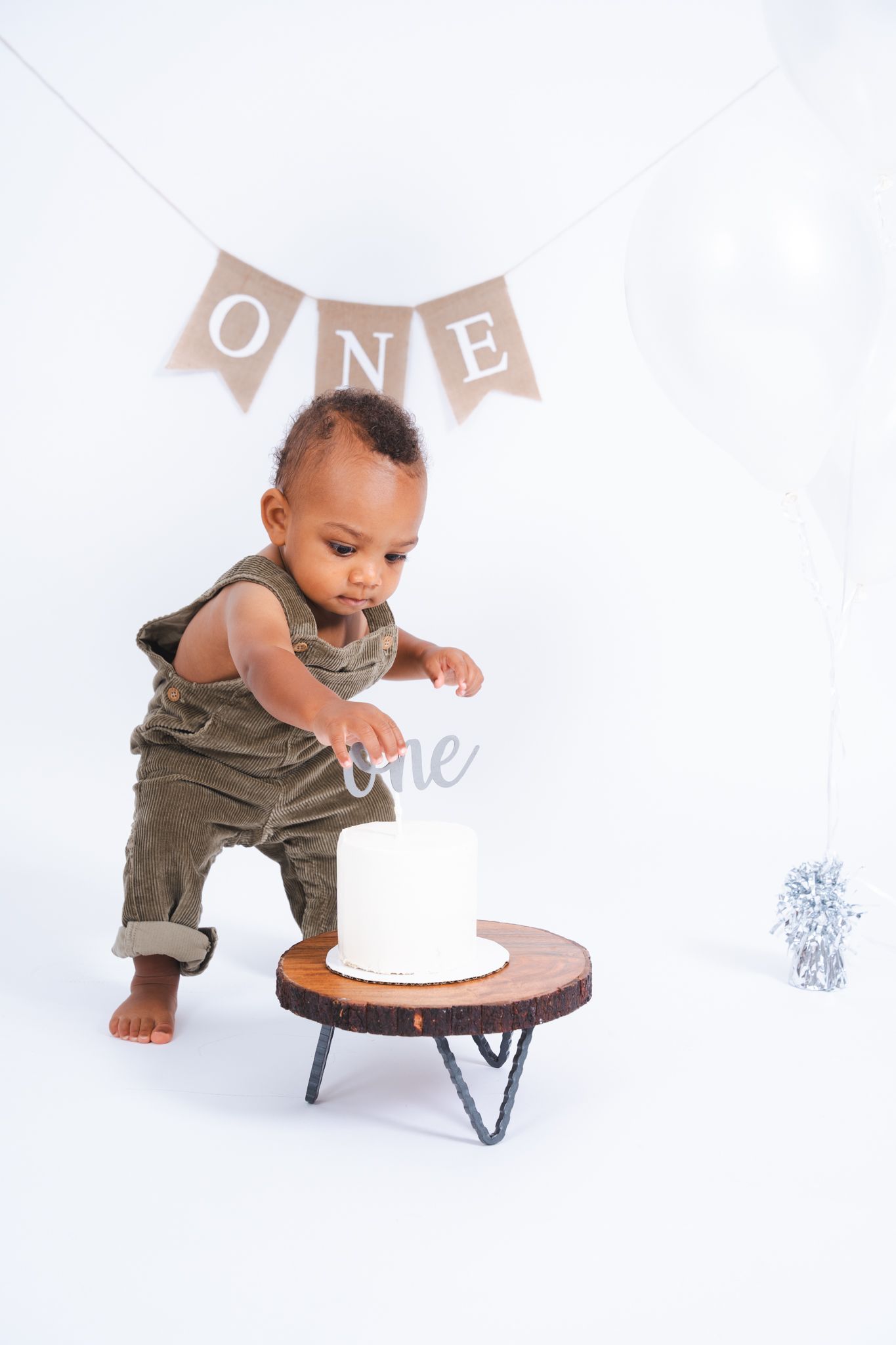 A small child in olive overalls reaches toward a white birthday cake on a wooden stand against a white backdrop.