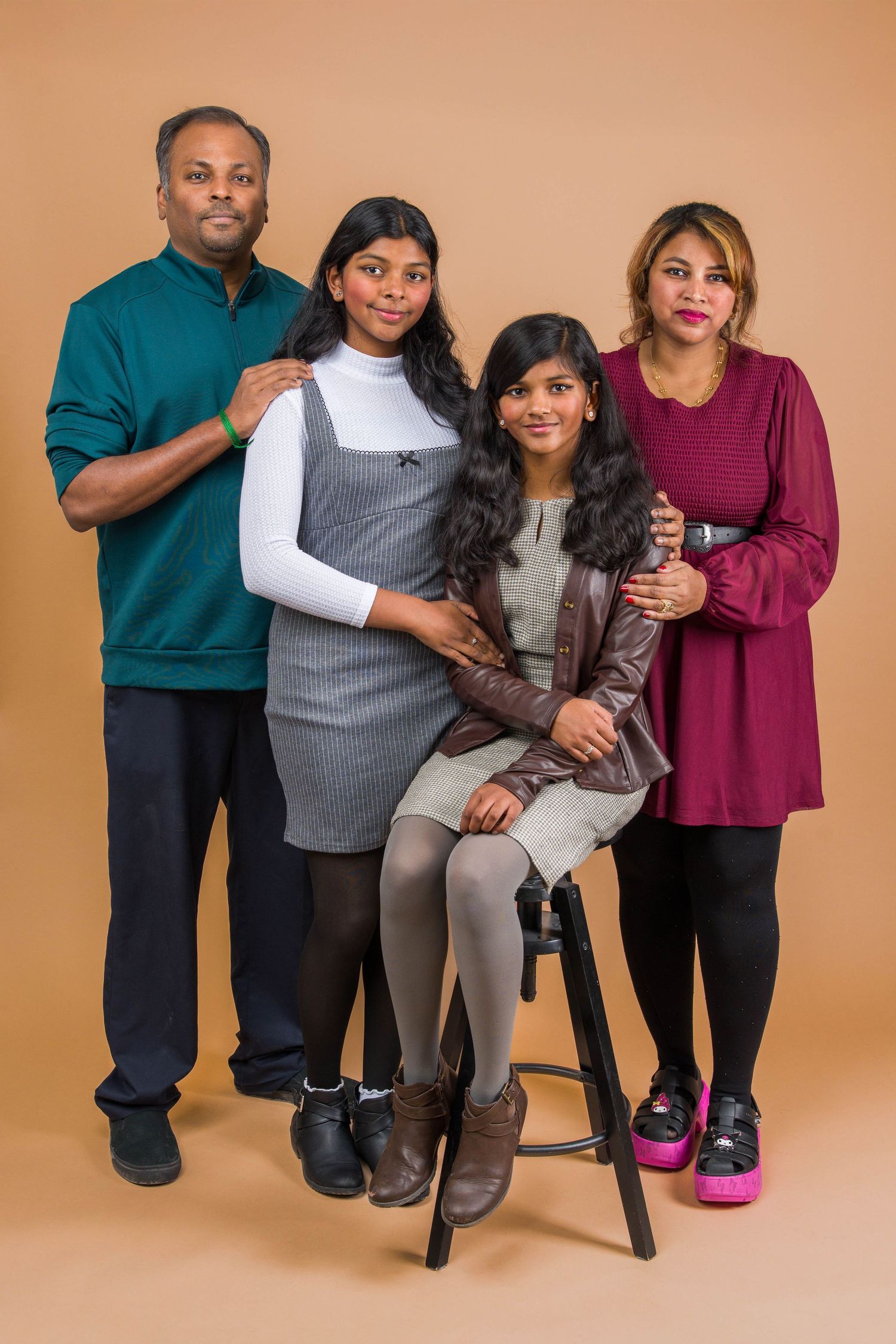 A family of four poses together for a studio portrait against a brown backdrop wearing coordinated casual outfits.