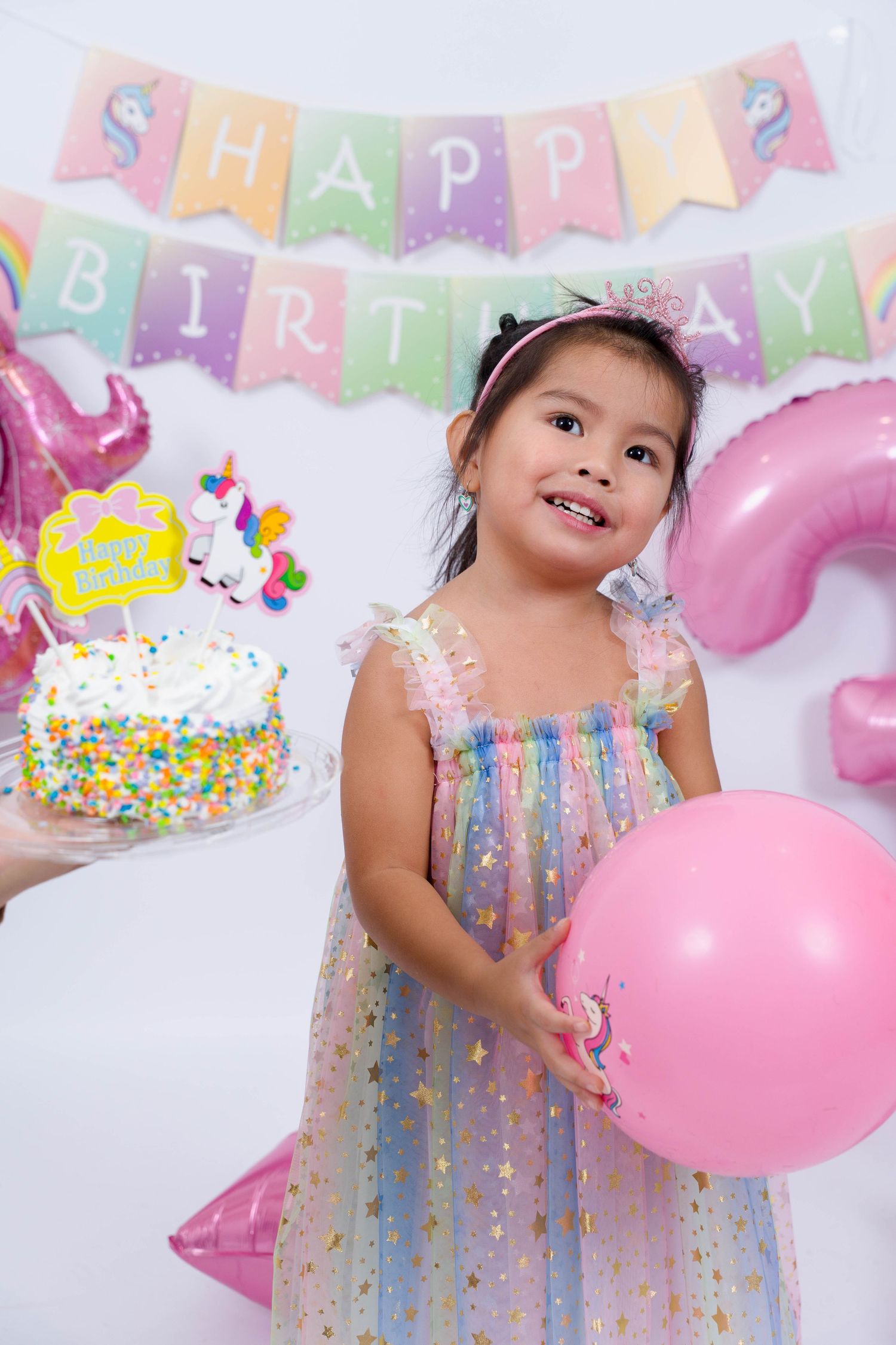 Colorful birthday party setup with pastel balloons, banners, and a decorated cake against a white backdrop.