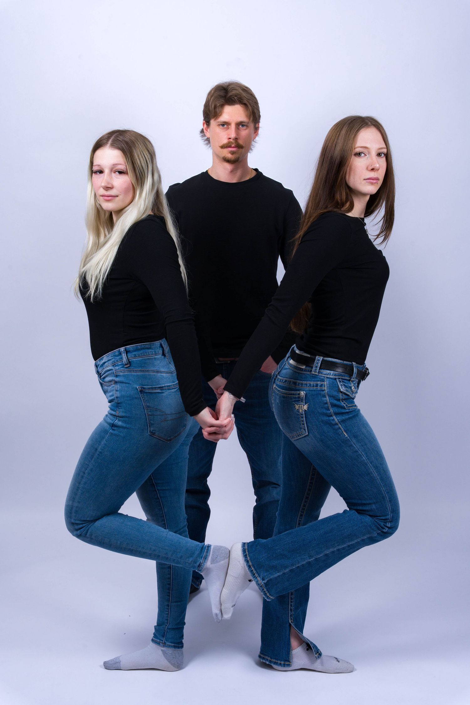 Three people in black tops and blue jeans pose in different positions against a white background for a fashion photo series.