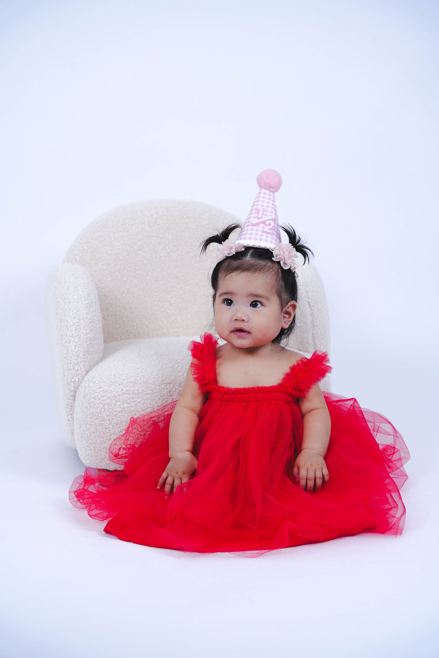 Small child in a red tulle dress and pink party hat sits on white armchair during photo session.