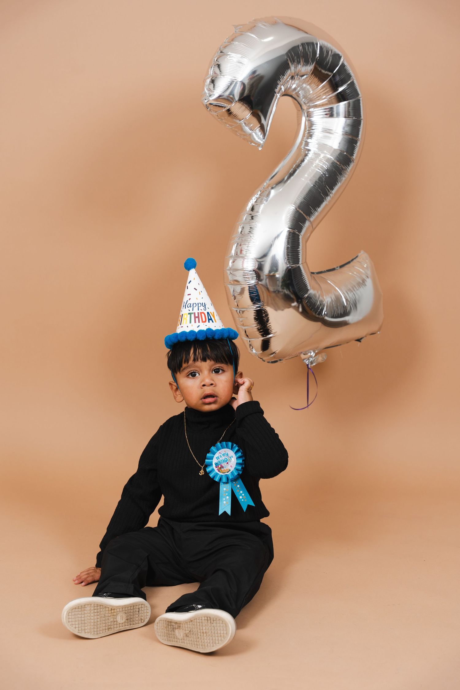 Child in black outfit sits on beige backdrop with silver number balloons for birthday photoshoot series.