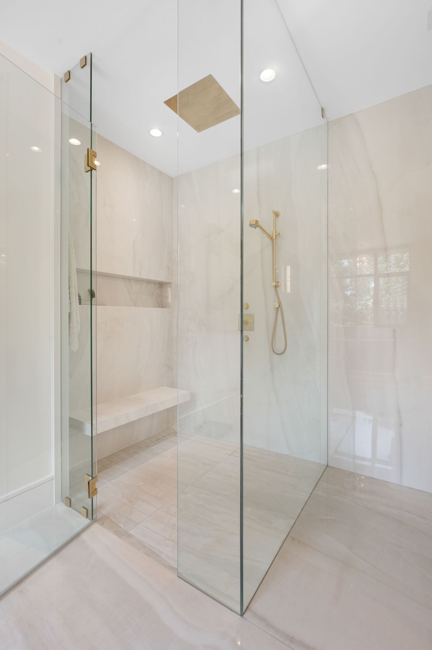 Minimalist walk-in shower with glass enclosure, light marble tiles, and gold fixtures.