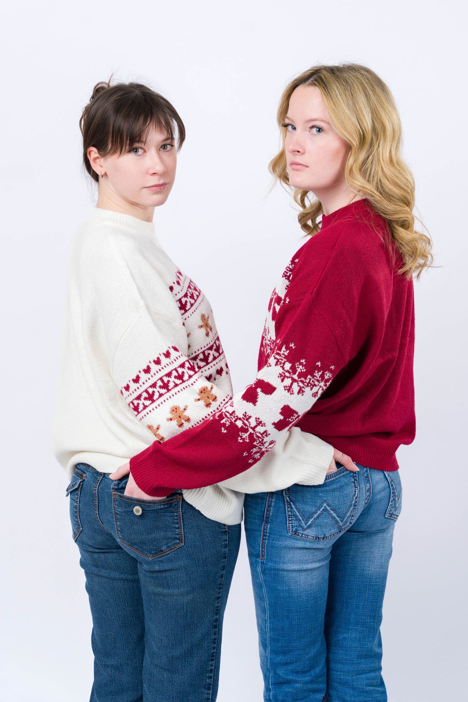 Two people stand back to back wearing matching winter sweaters with Nordic patterns in red and white with blue jeans.