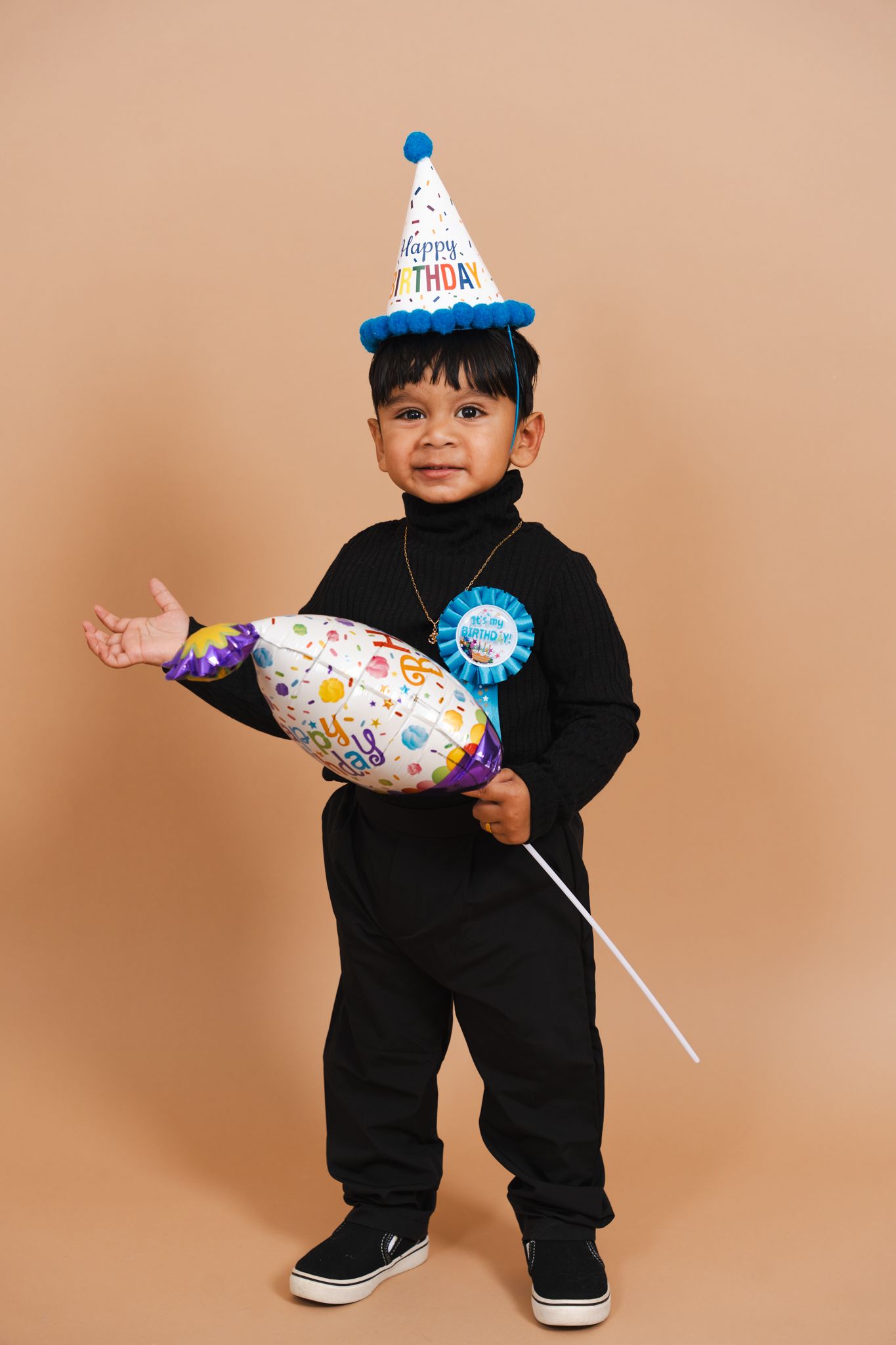 A small child wearing a black outfit and birthday party hat holds a festive balloon against a beige background.