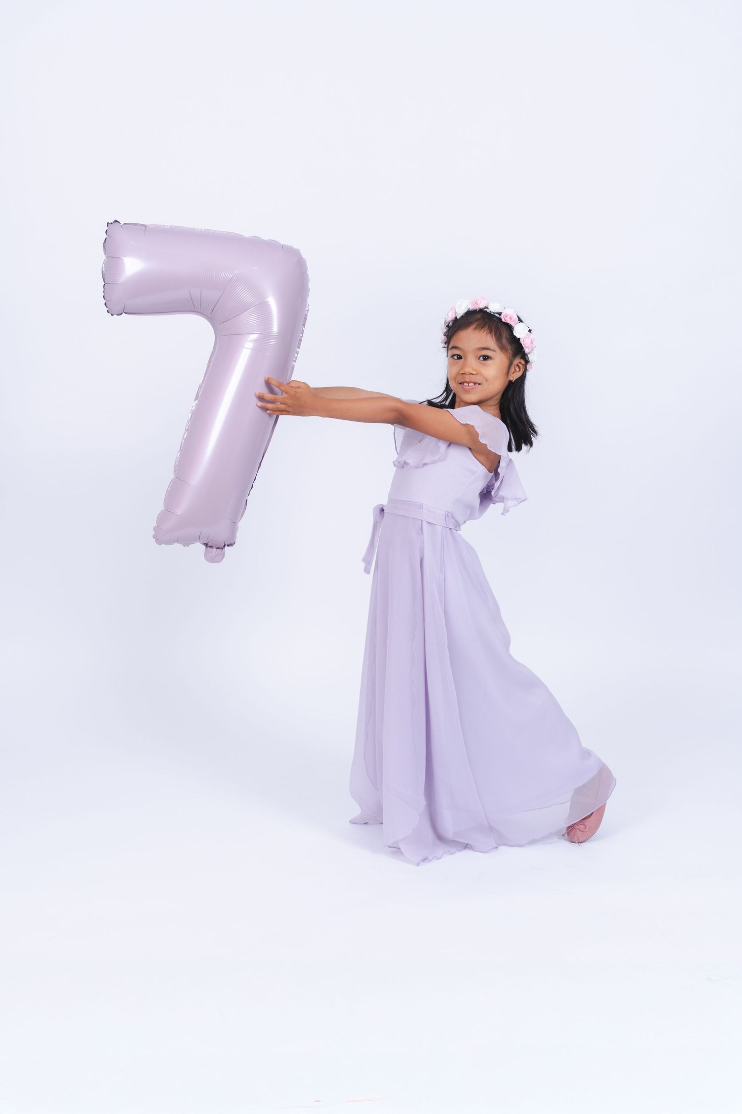 A young child in a lavender dress poses with a large number 7 balloon against a white background.
