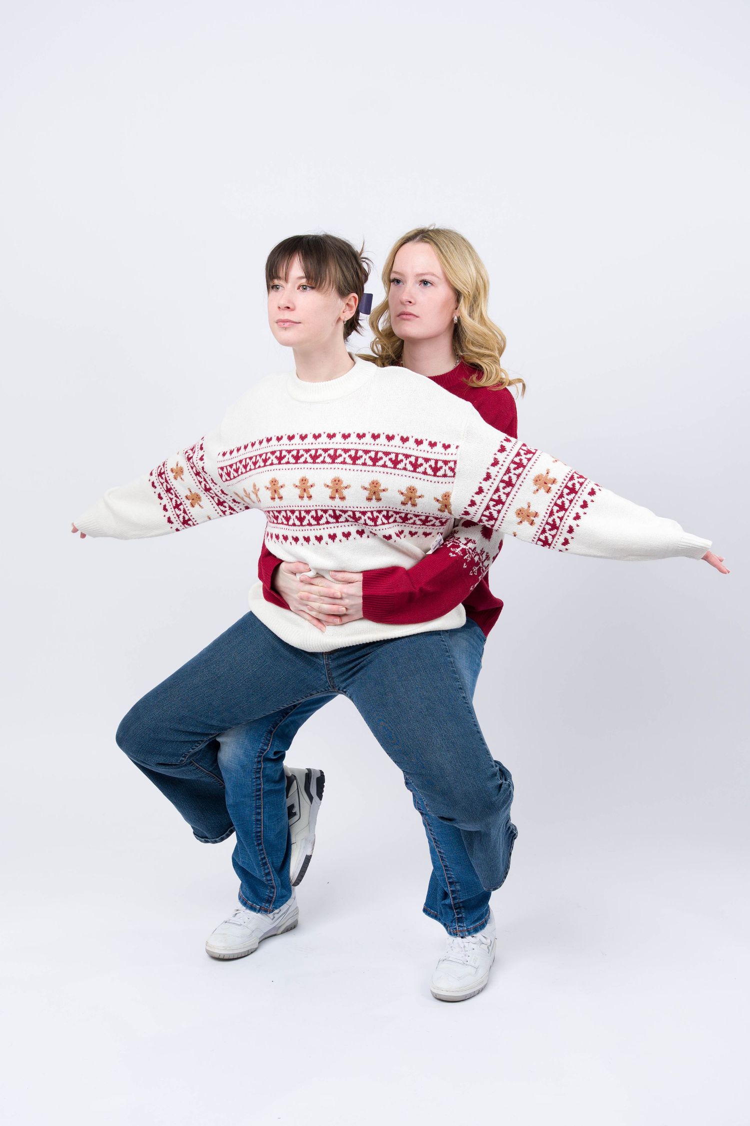 Two people in matching holiday sweaters and jeans strike a playful pose against a white background.