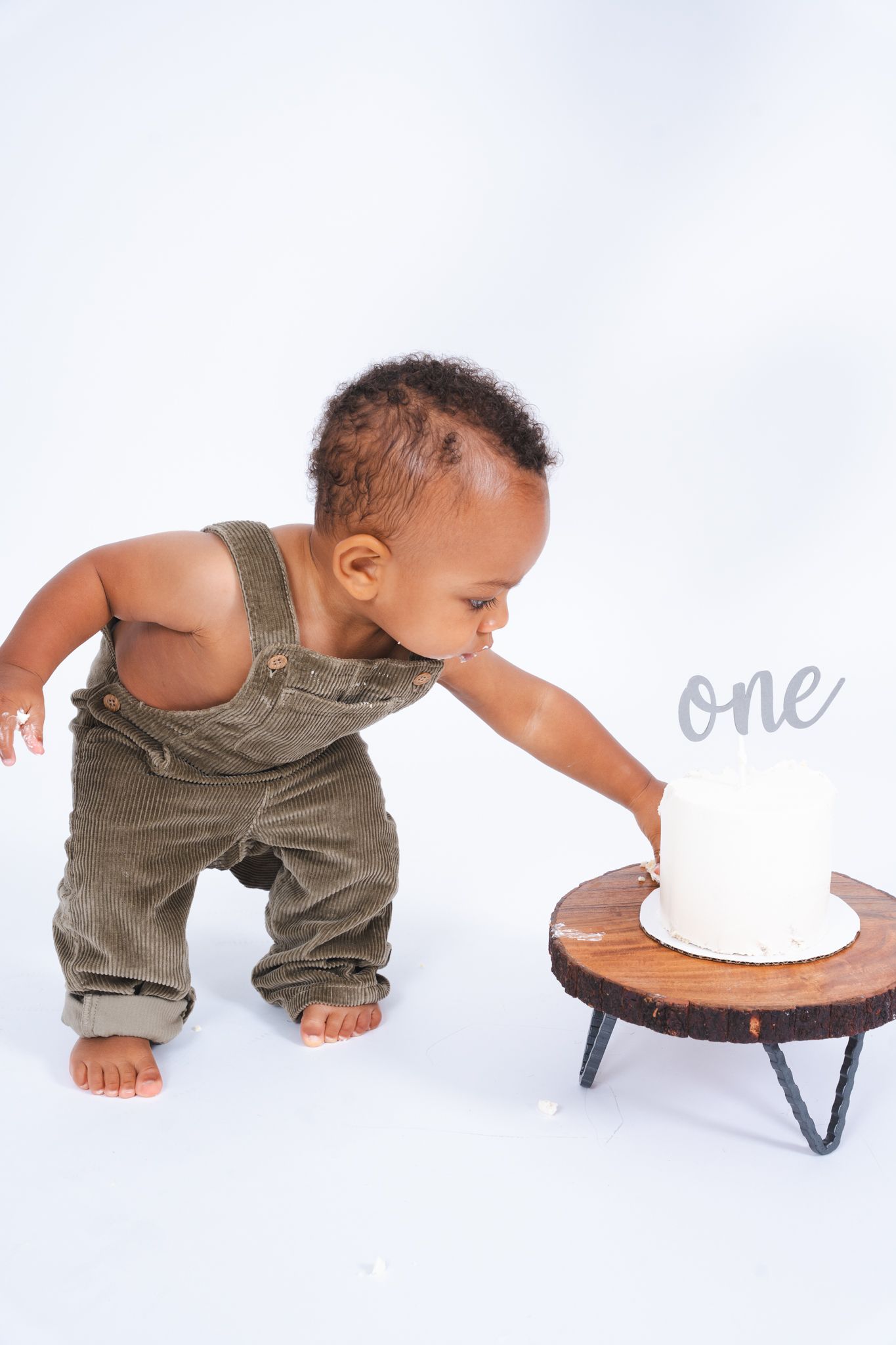 Baby in olive green overalls reaches toward a simple white birthday cake with 'one' text on a wooden stand.