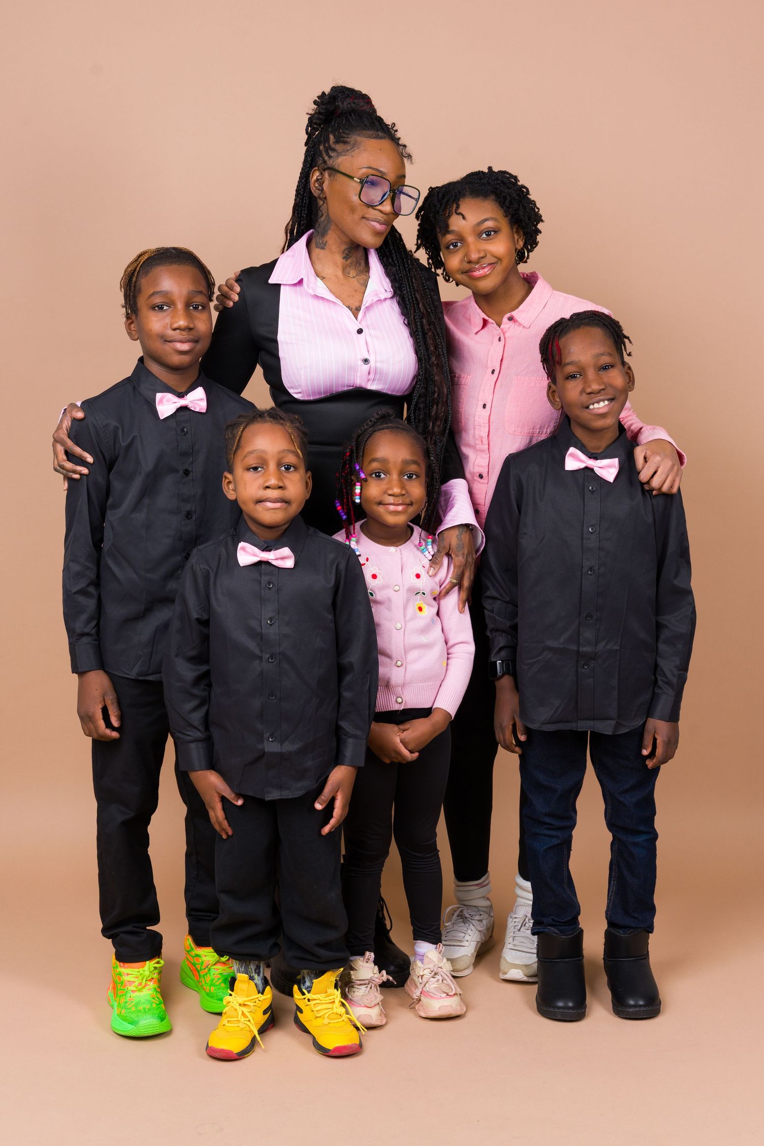 Group of children wearing matching black jackets and colorful sneakers pose together against pink backdrop.