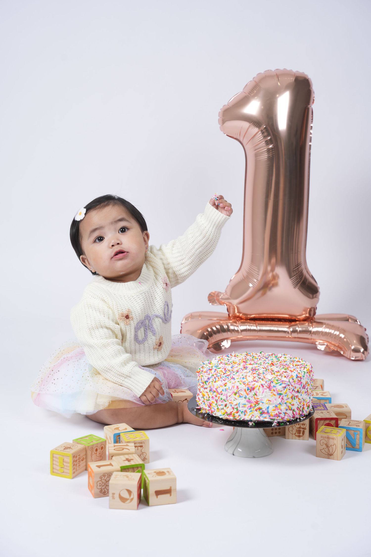 Baby in cream sweater plays with wooden blocks and rose gold balloon number one in first birthday photo series.