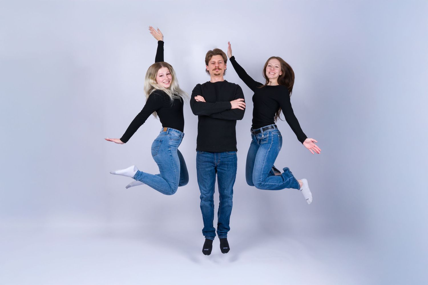 Three people jumping in sync against a gray background wearing casual clothing and jeans.