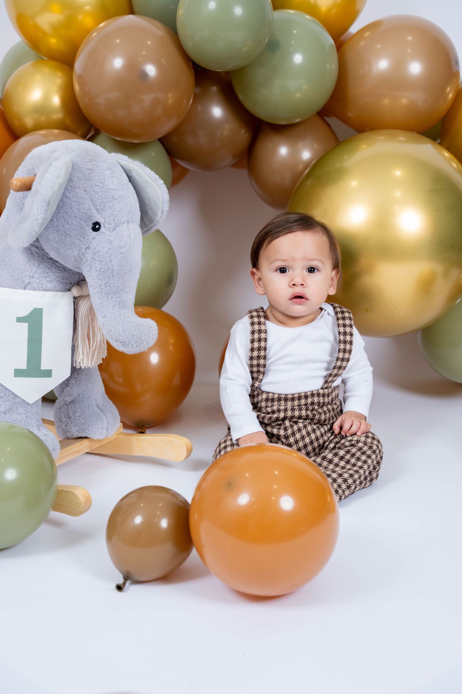 Baby's first birthday party setup with gold and sage green balloon garland against white backdrop.