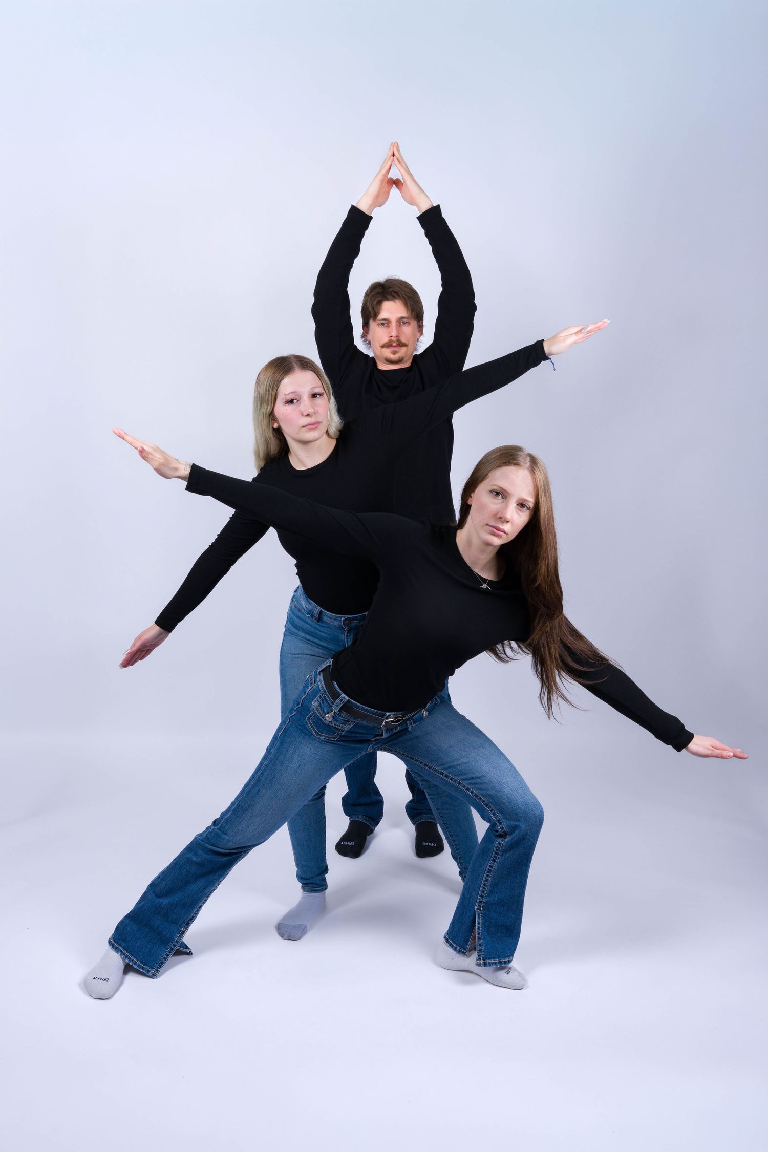 Three performers in black tops and jeans strike a dynamic pose with outstretched arms against a light background.