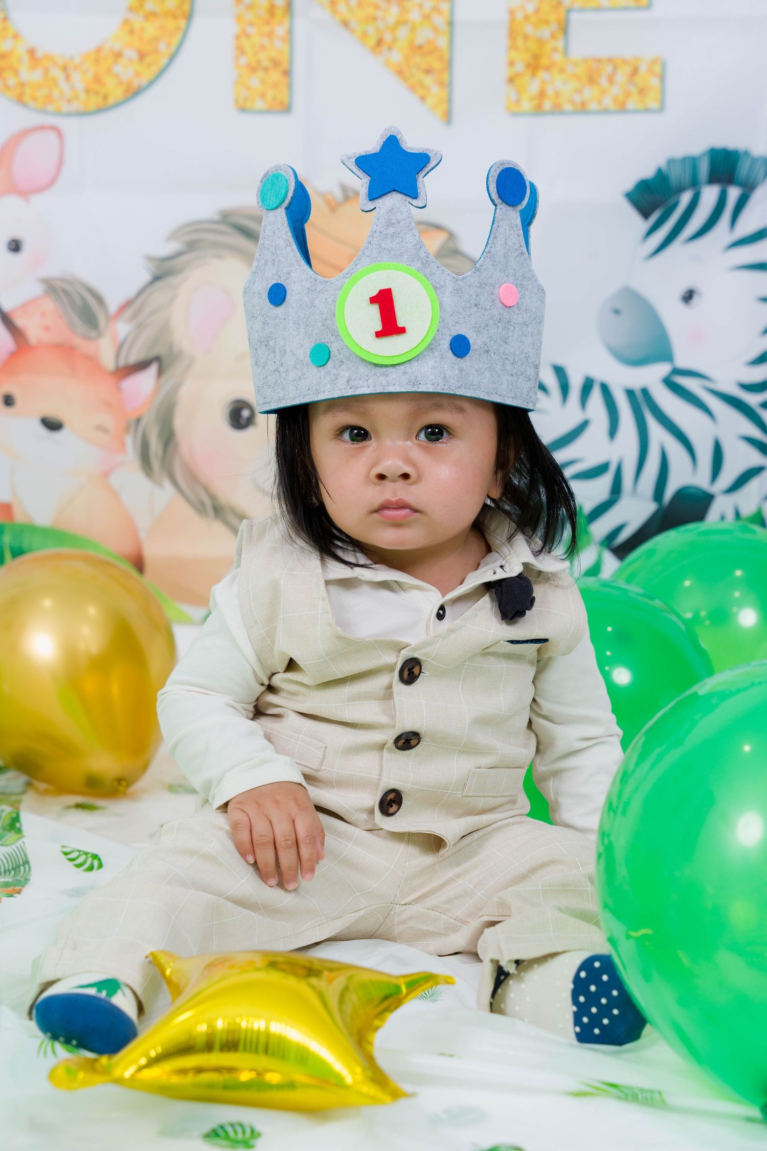A baby in a beige outfit sits among colorful balloons and tropical leaf decorations at a birthday celebration.
