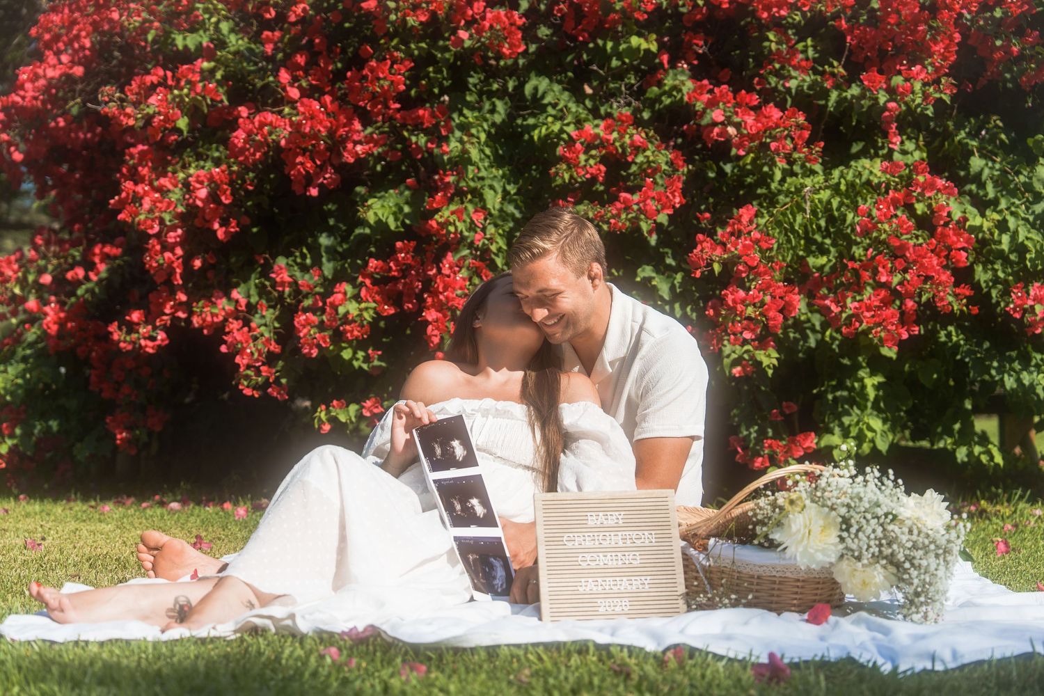 girl kissing man in front of red flowers in a dreamy baby announcement photoshoot