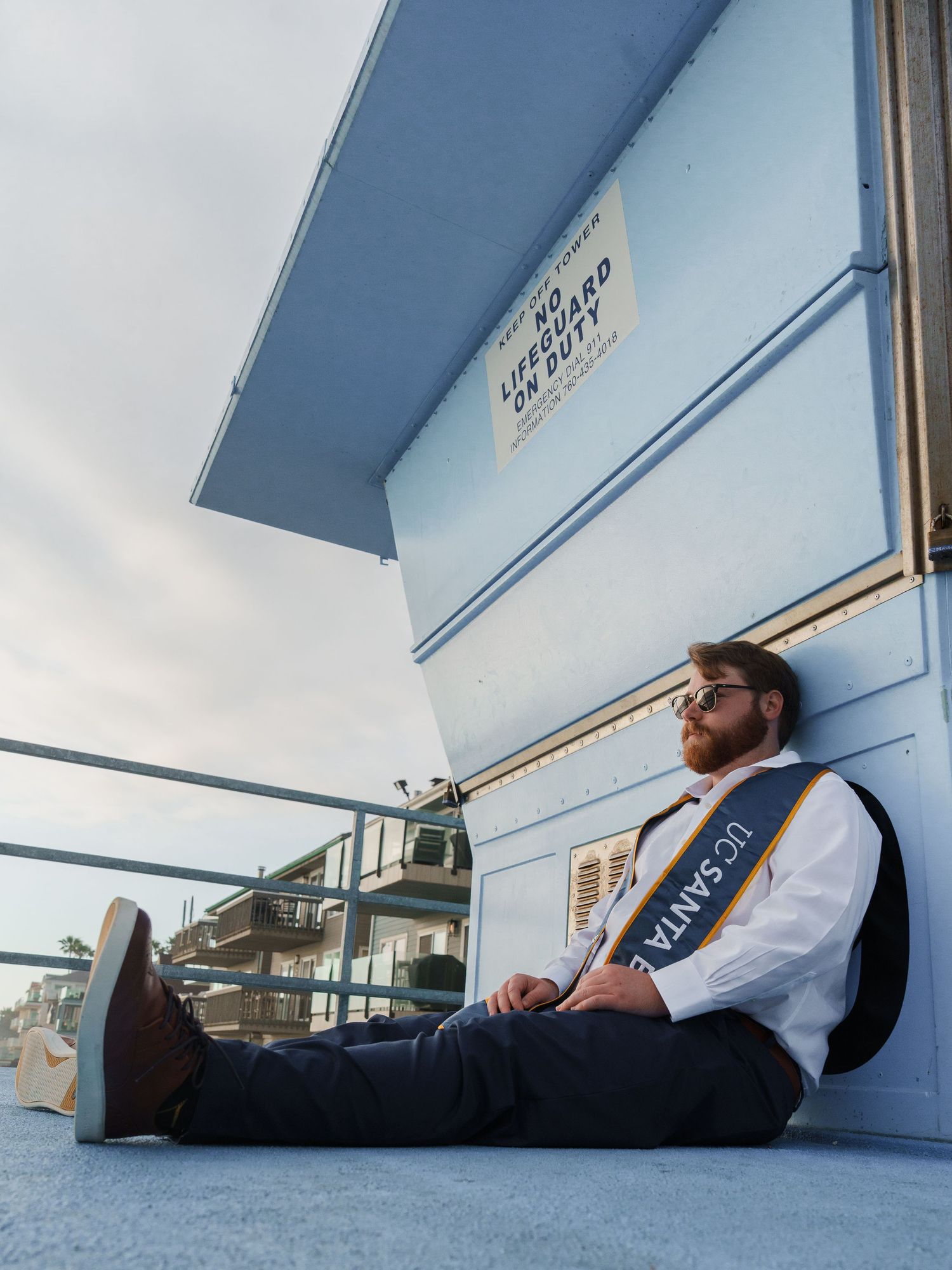 Someone in a vest and dress shirt sits casually against a light blue wall of a building under an overhanging roof with a UCSB stole on.