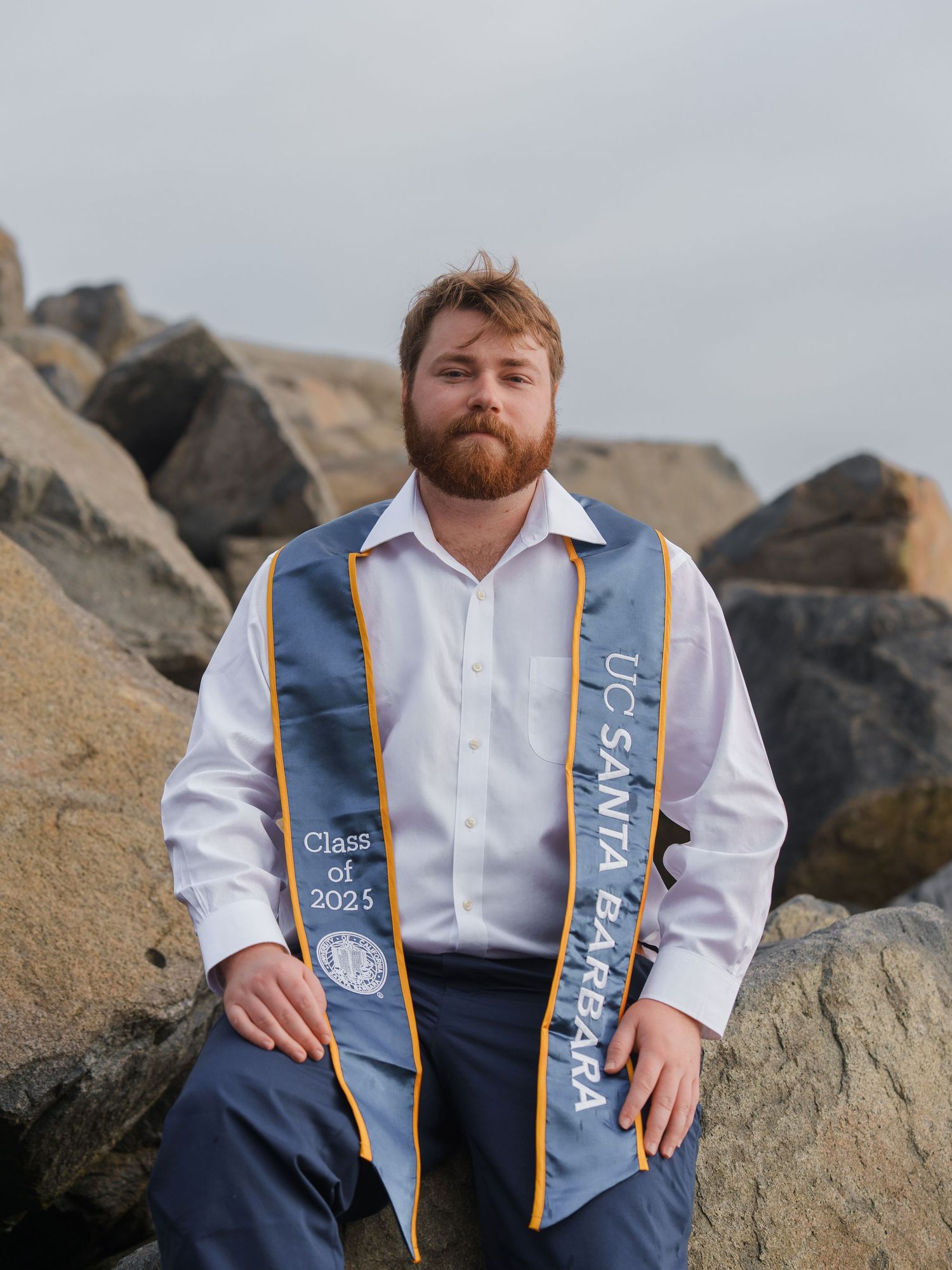 man posing for a graduation photo with a UCSB stole on his shoulder, at the beach, while sitting on a rock