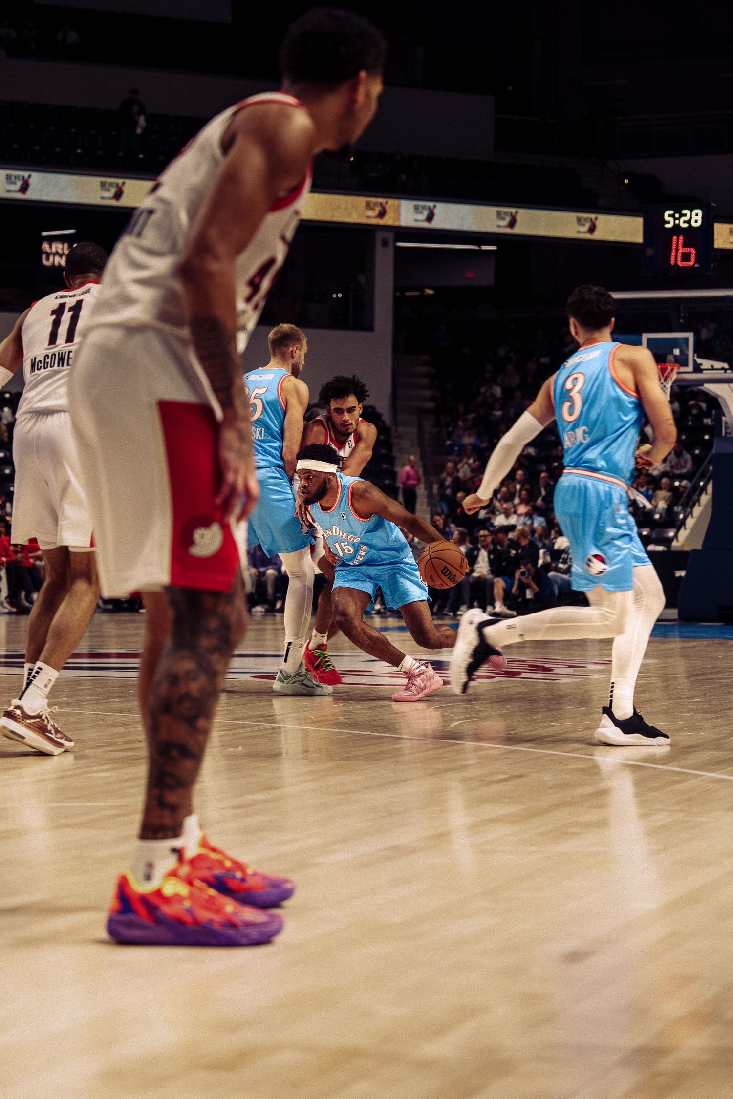 Basketball players in light blue and red uniforms move across a wooden court during a game.