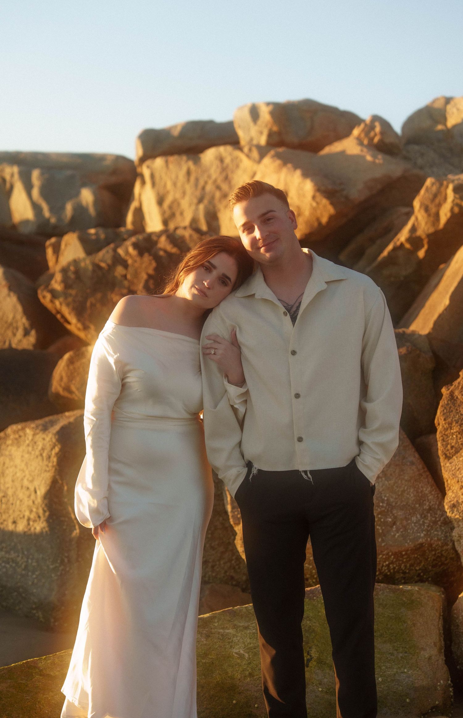 A couple in matching cream-colored outfits pose together against golden rocks at sunset.