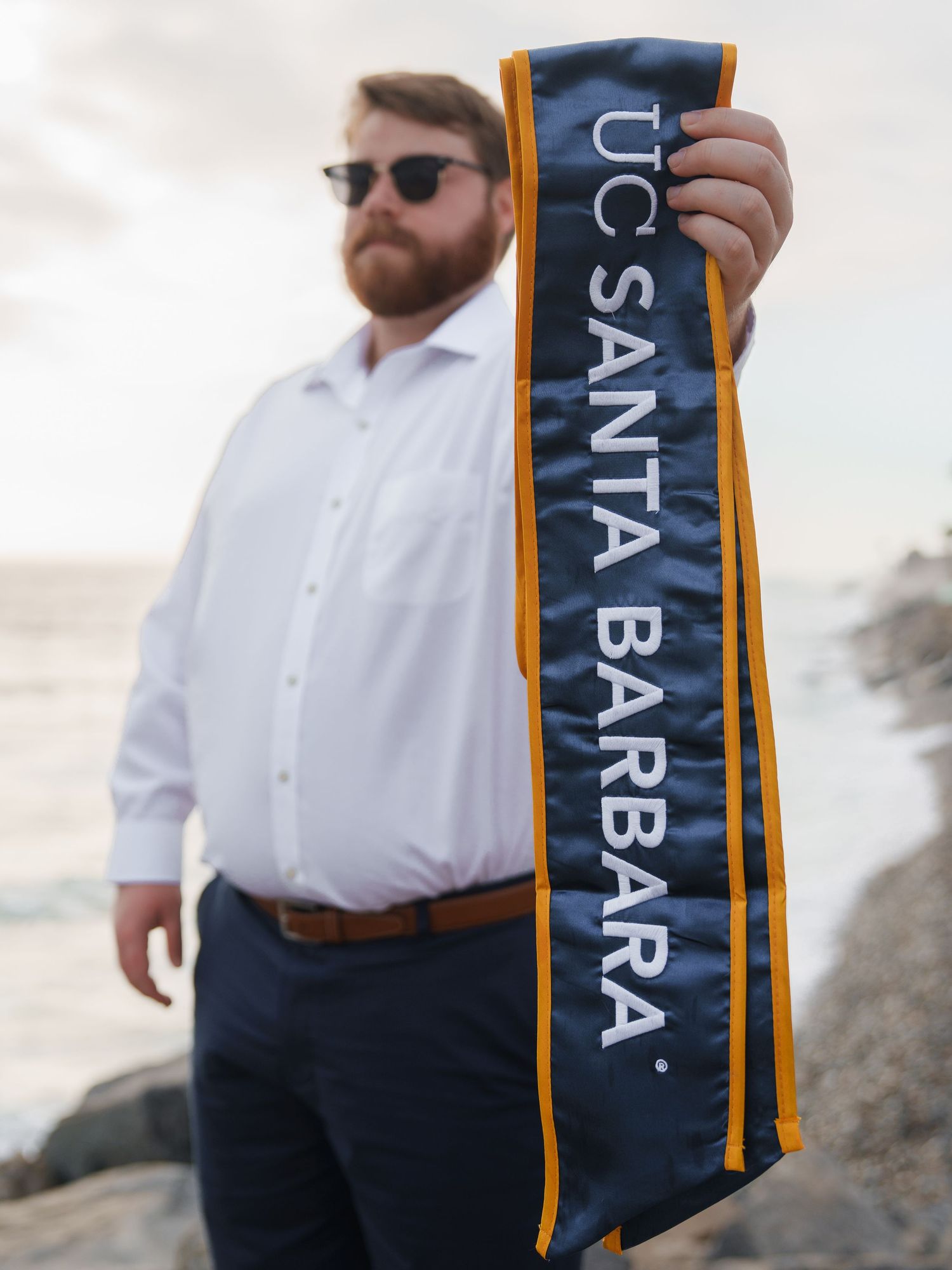 man holding his USCB graduation stole while standing on a rock at the beach