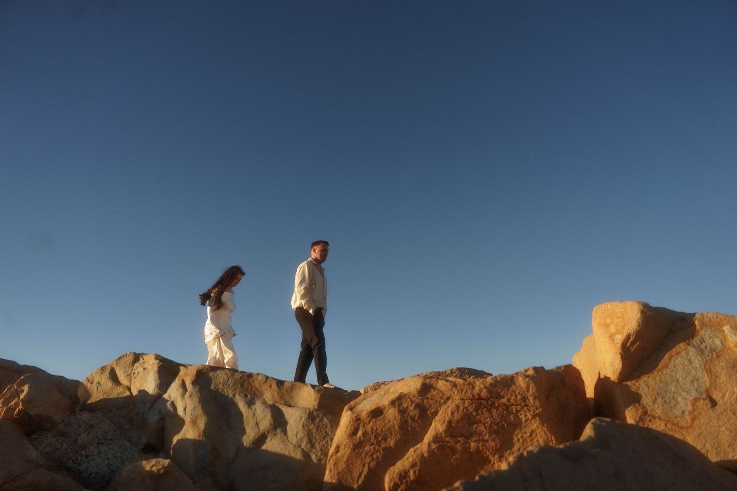 Two people walking on large rocky boulders against a blue sky at sunset.