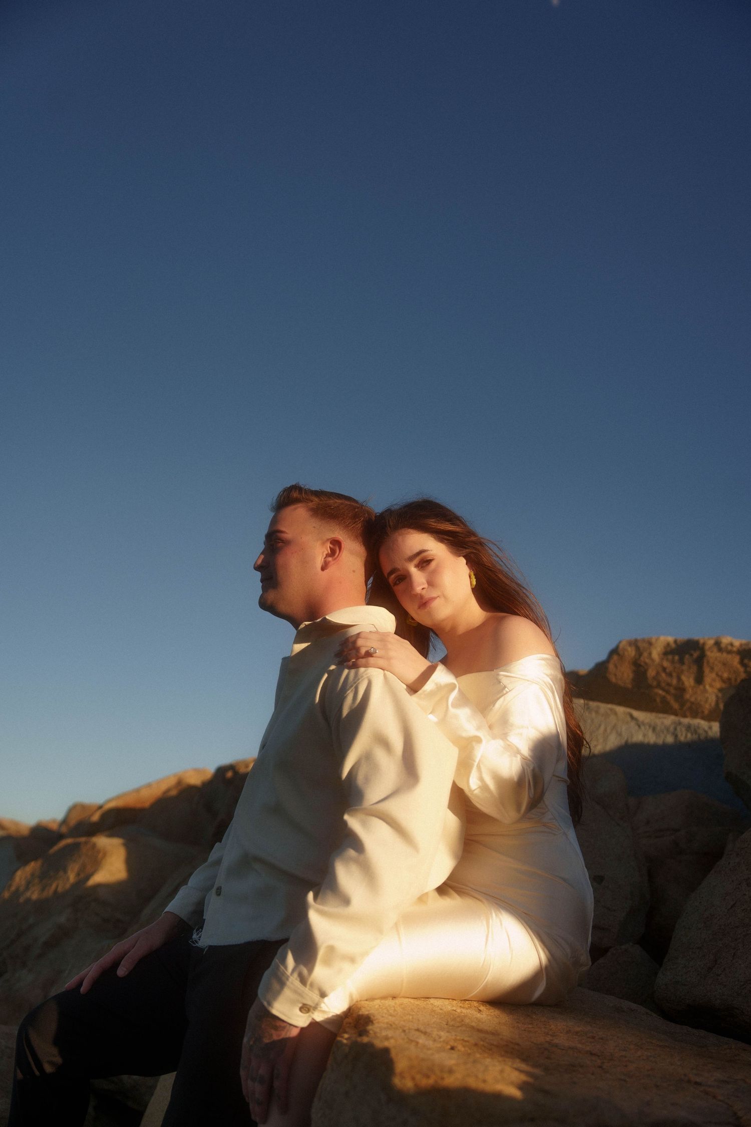 A couple in white outfits sits together on desert rocks at sunset against a blue sky.
