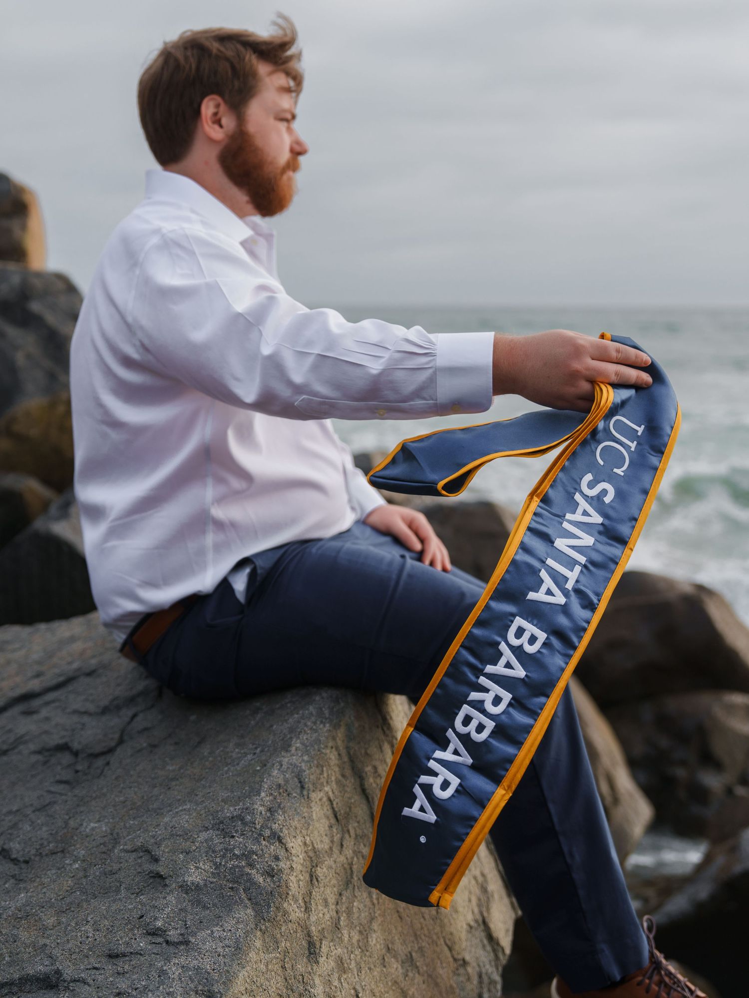 man holding his USCB graduation stole while sitting on a rock at the beach