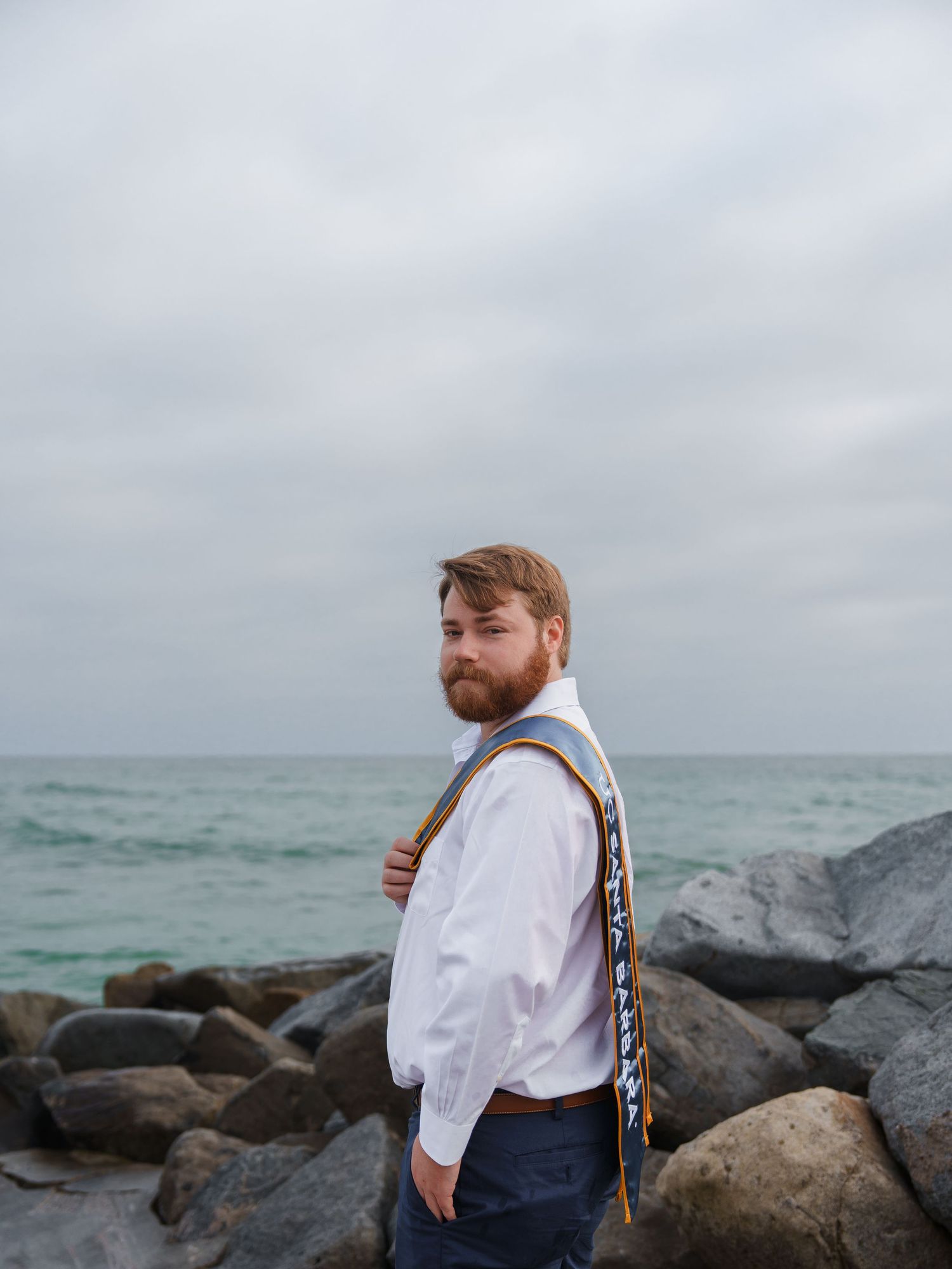 man posing for a graduation photo with a UCSB stole on his shoulder, at the beach