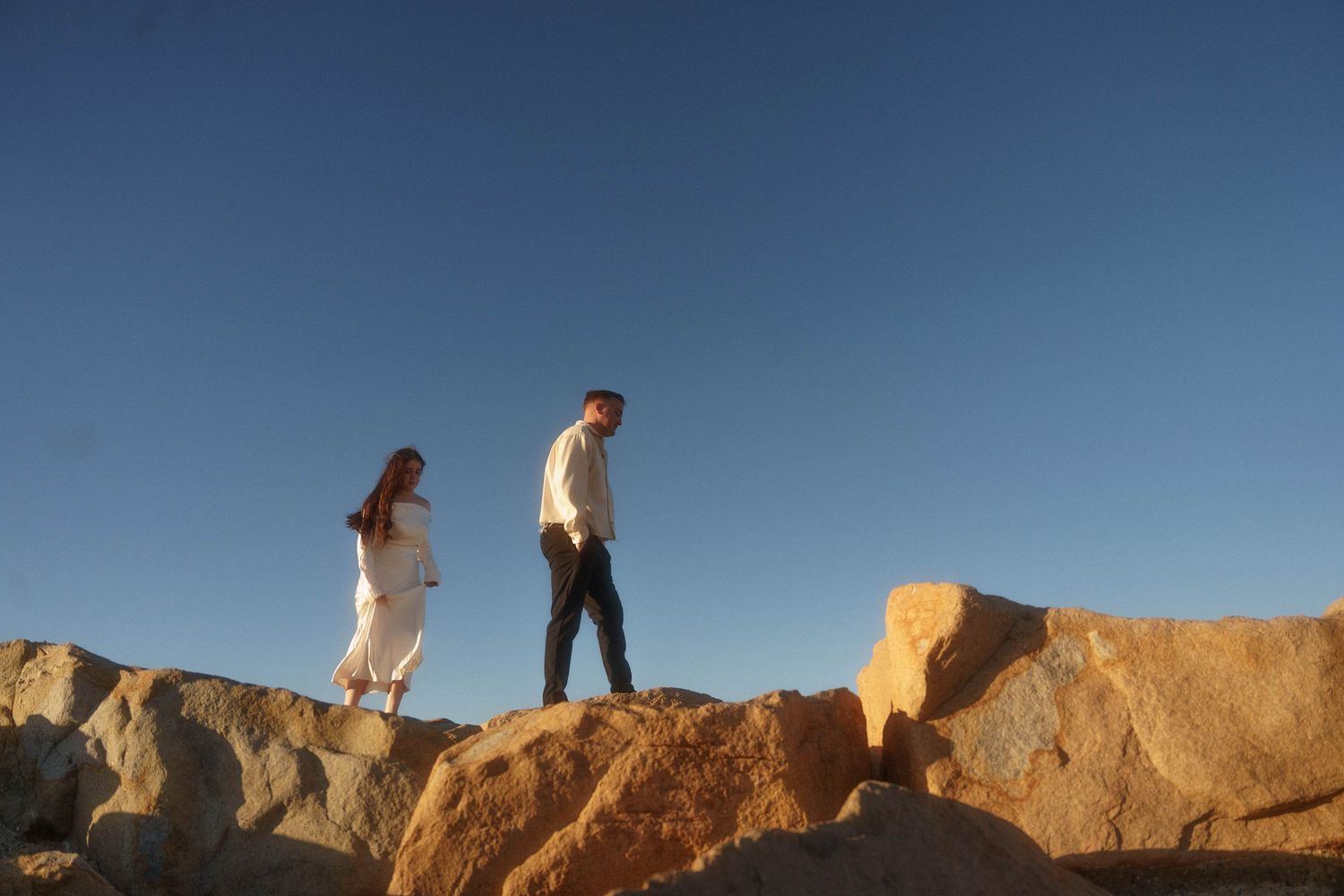 Two people stand on a rocky cliff edge overlooking a scenic desert landscape at sunset.