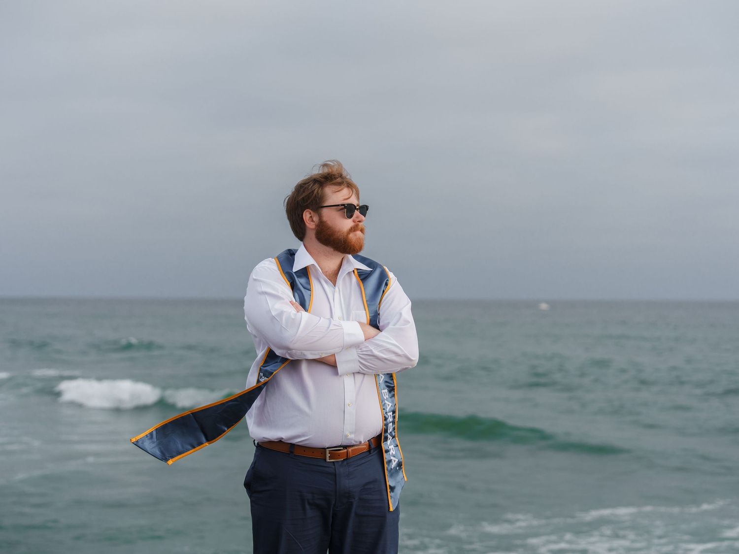 man posing for a graduation photo with a UCSB stole on his shoulder, at the beach