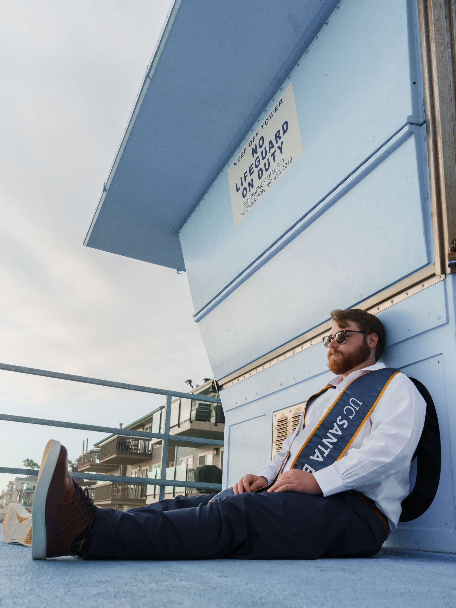 man sitting at a lifeguard tower with a UCSB stole on his shoulder staring in the distance for a graduation photo