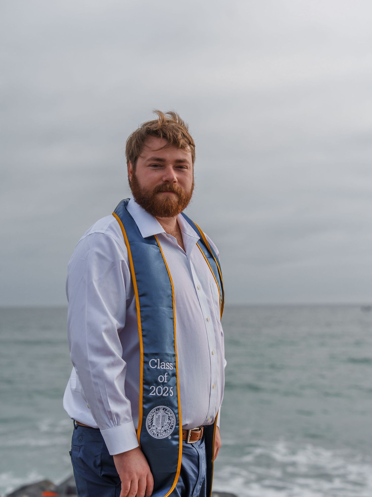 man posing for a graduation photo with a UCSB stole on his shoulder, at the beach