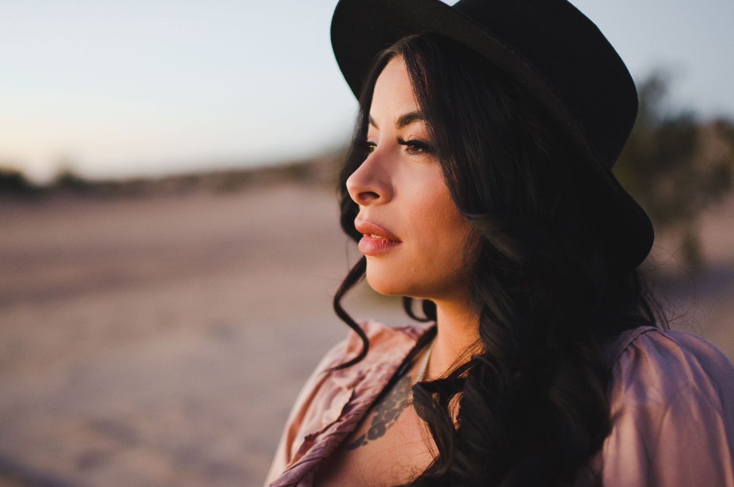 Beautiful woman with dark curly hair and floral tattoo wearing black hat at sunset.