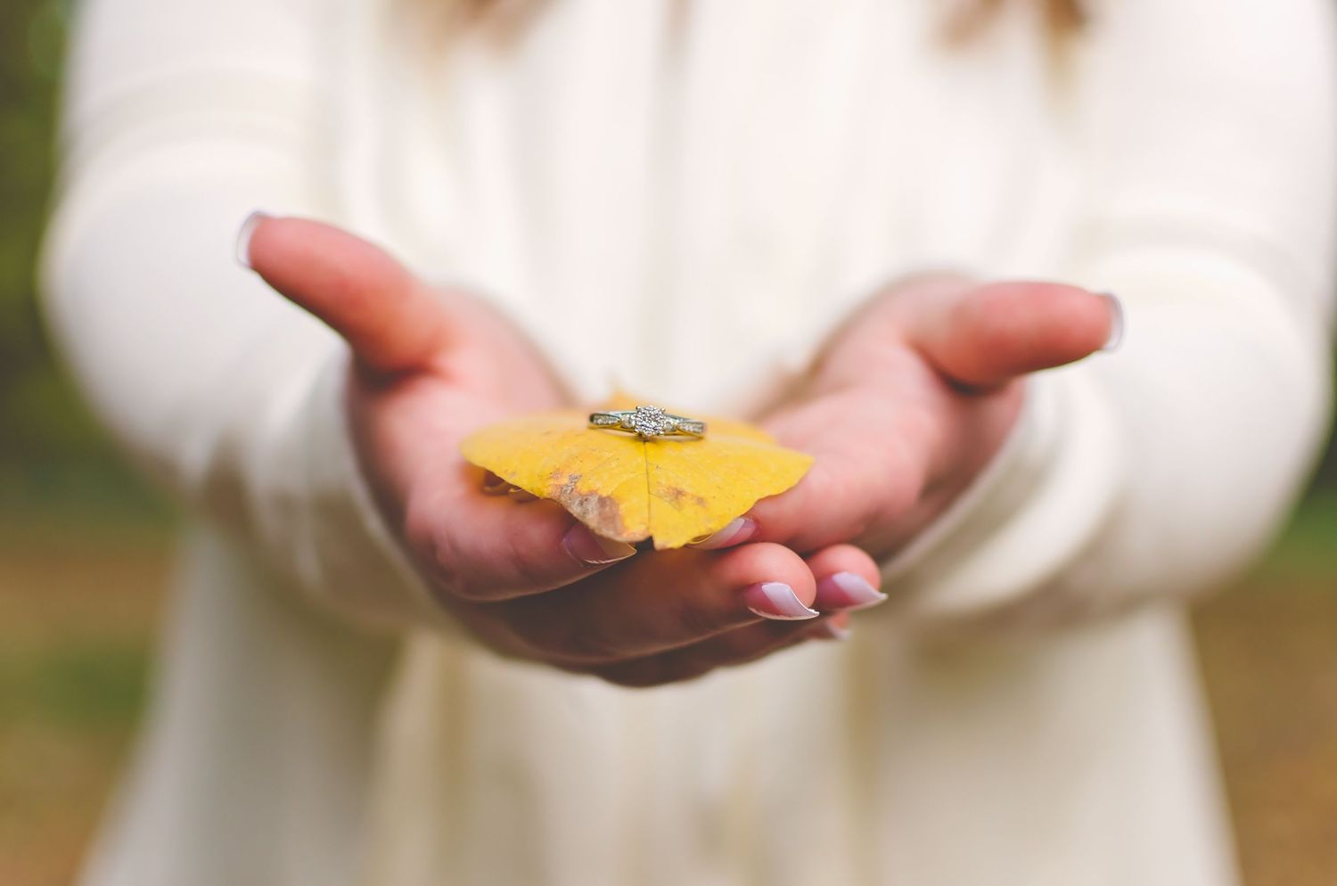 Close-up of woman in white sweater holding yellow ginkgo leaf with small ring resting on top.